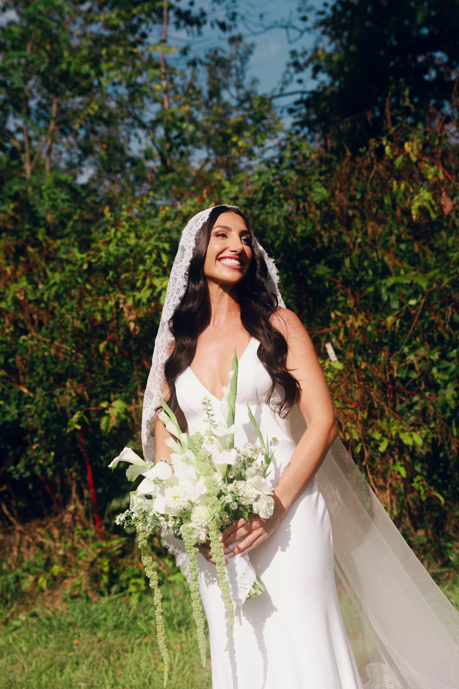 Smiling bride in white gown and lace veil holds white floral bouquet outdoors with lush green trees behind her.