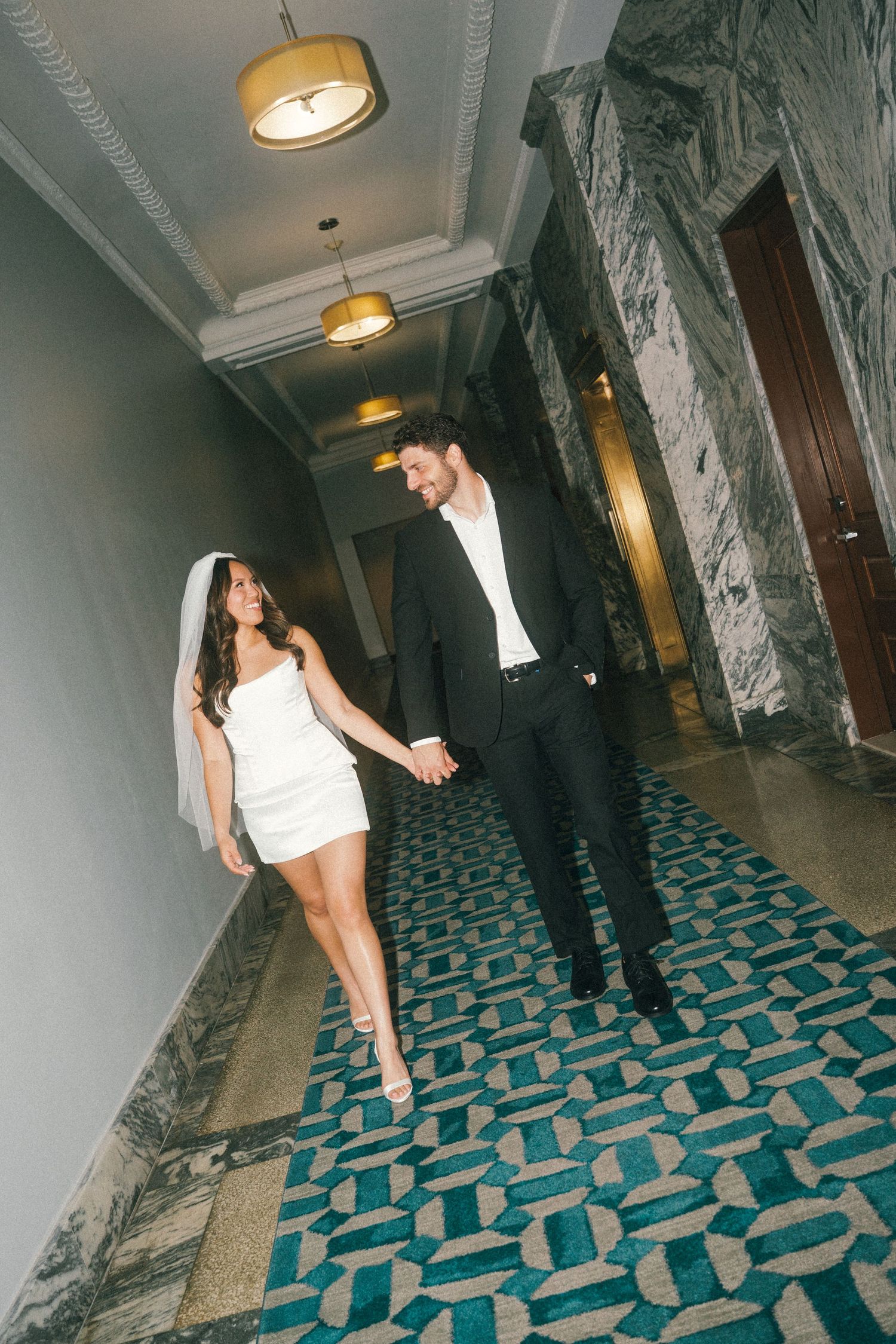 A couple walks playfully down a hallway with distinctive teal and white geometric carpet.