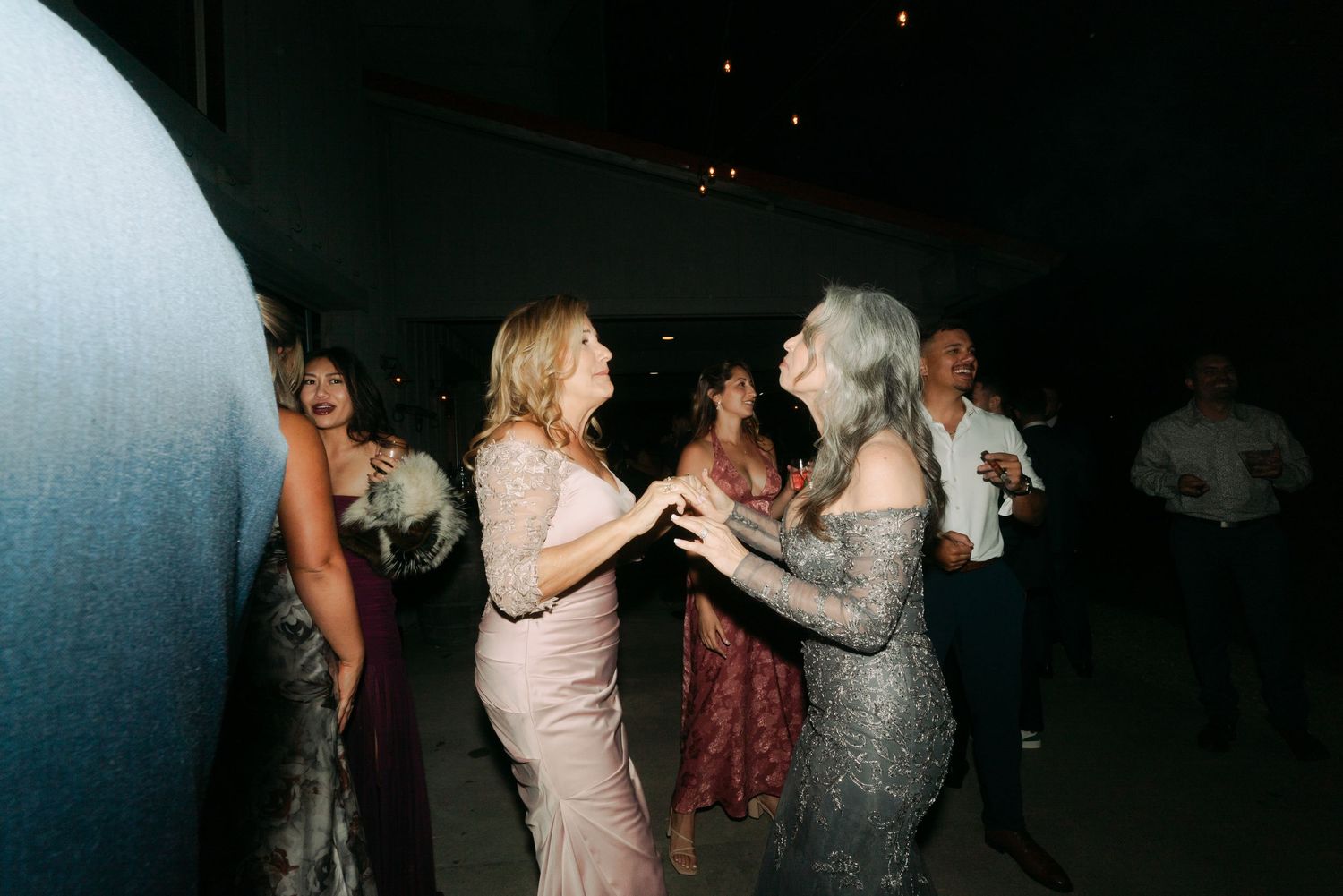 Women in elegant gowns dancing together joyfully on the dark wedding reception dance floor.