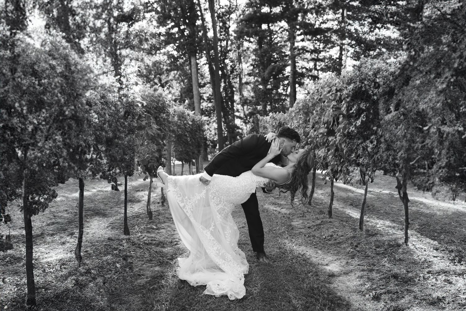 A romantic wedding couple dips and dances together in a wooded outdoor setting captured in black and white.