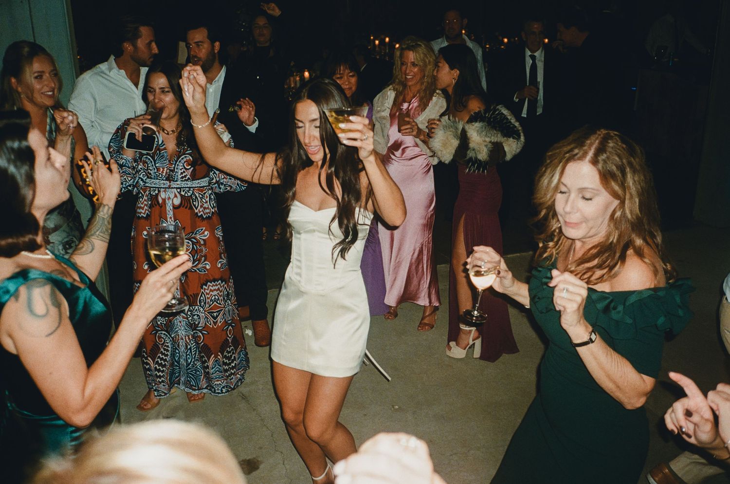 Bride in white mini dress dancing joyfully with guests at lively wedding reception celebration.