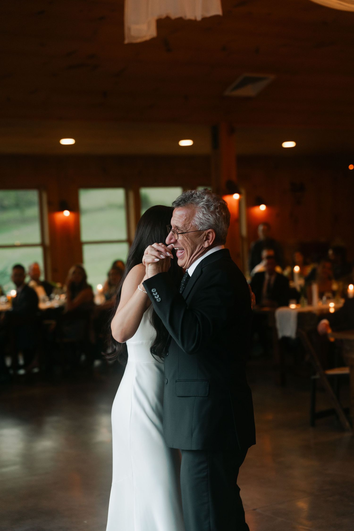 Bride shares an emotional father-daughter dance in a warmly lit rustic wedding reception venue.