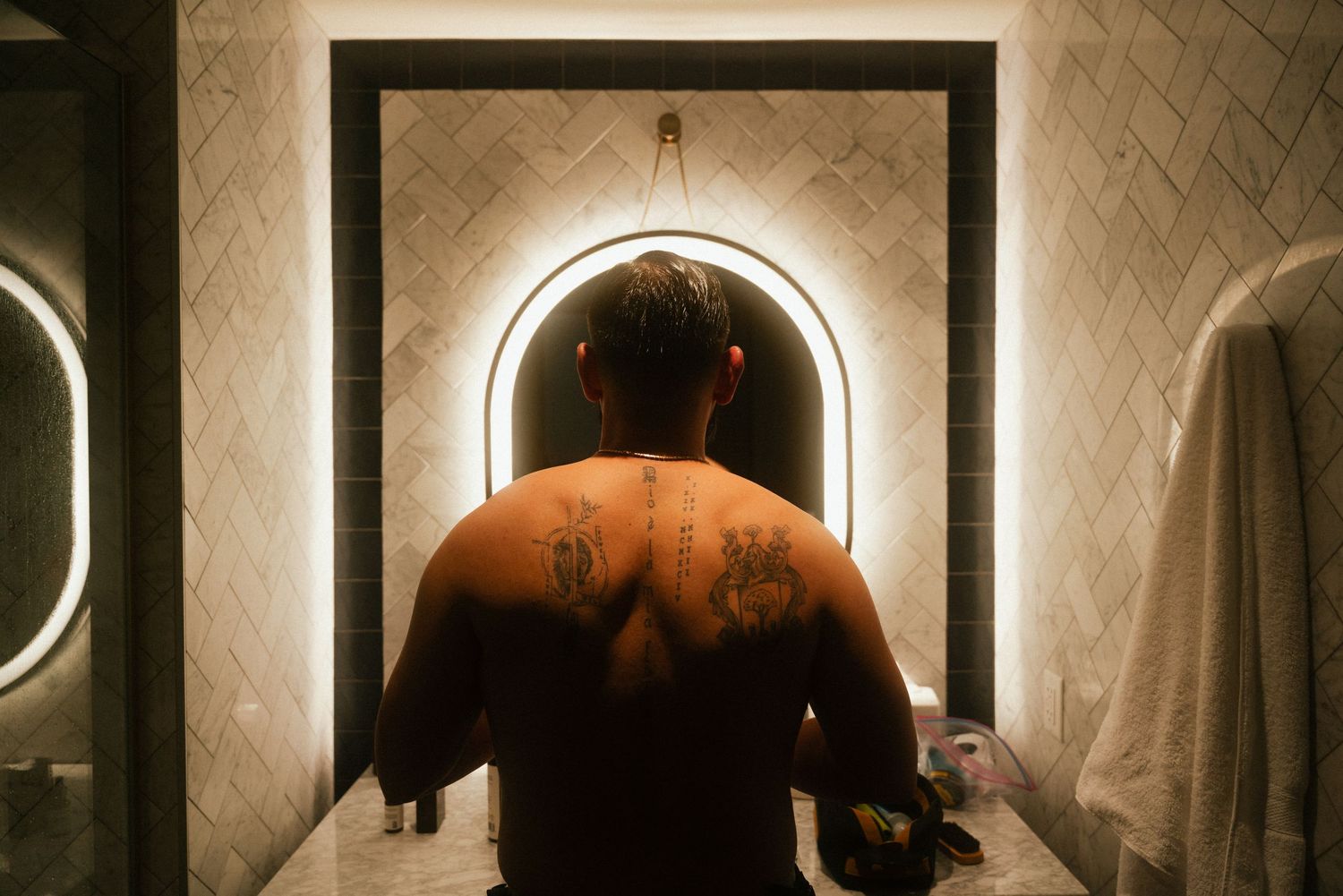 Man with back tattoos standing shirtless in front of illuminated arched bathroom mirror.
