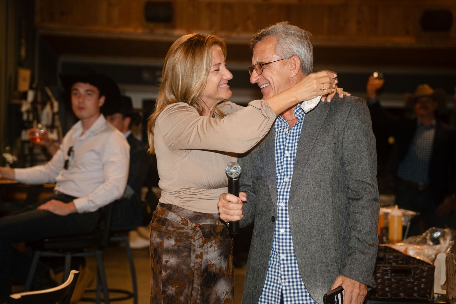 A woman and man share a warm embrace at a social gathering, with guests seated in the background.