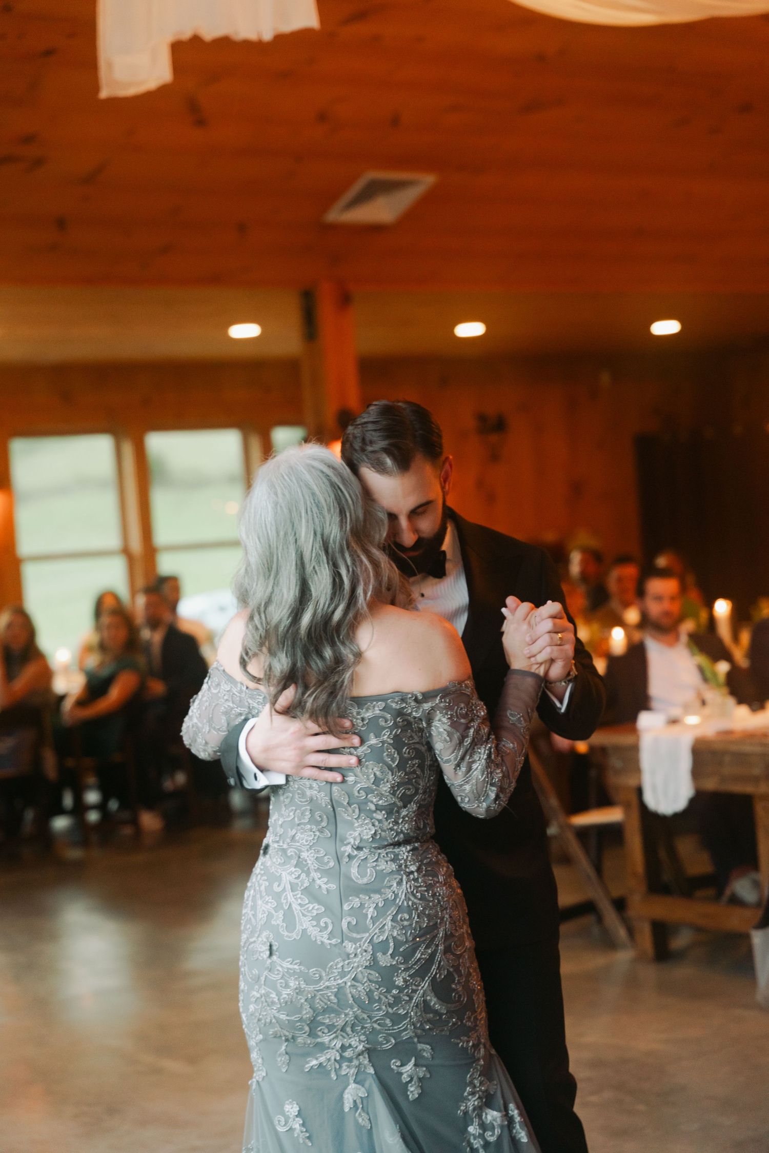 Groom shares a tender mother-son dance with his mother in a stunning lace gown at a rustic reception.