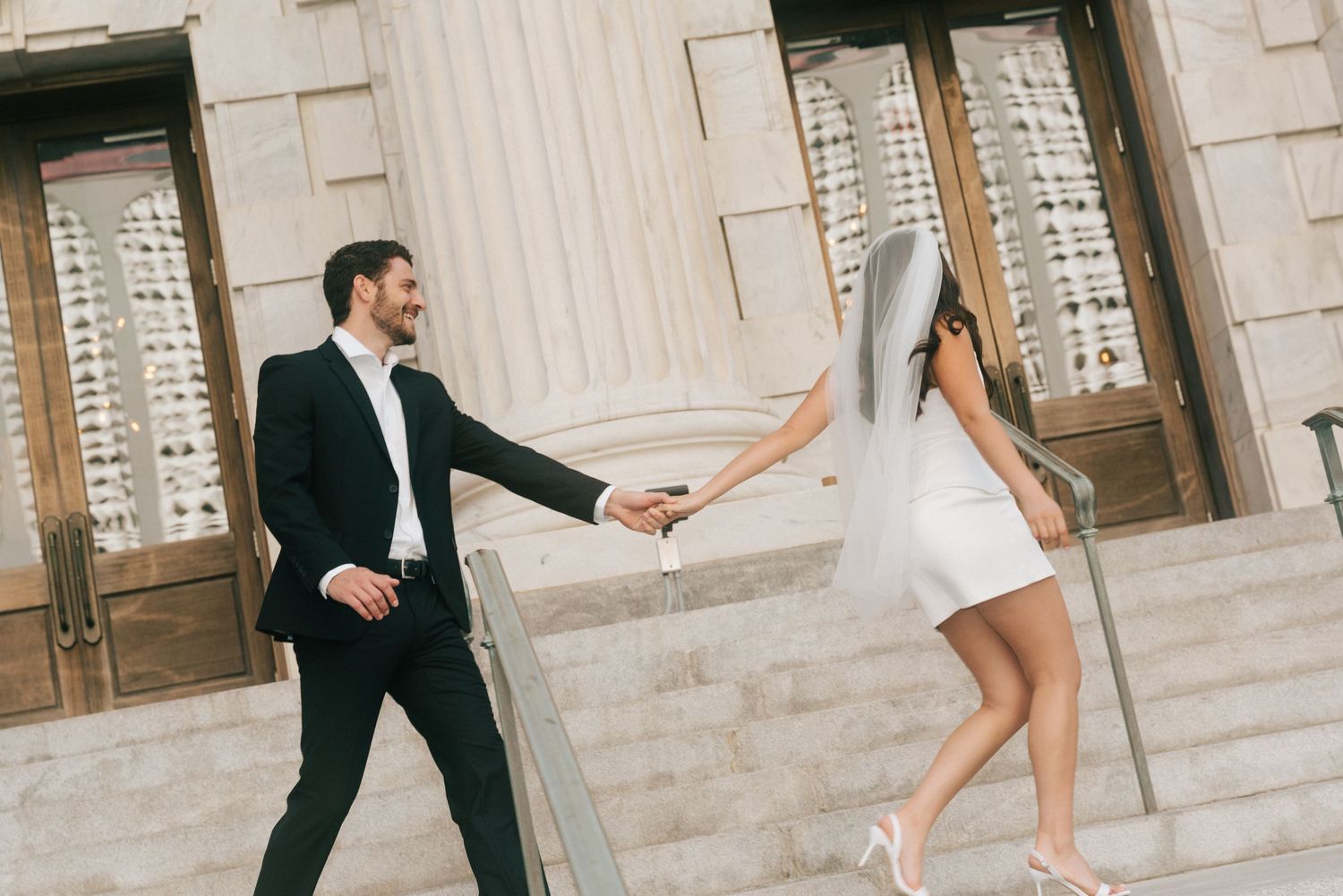 Bride and groom holding hands on stone steps, woman in white mini dress and veil, man in black suit.