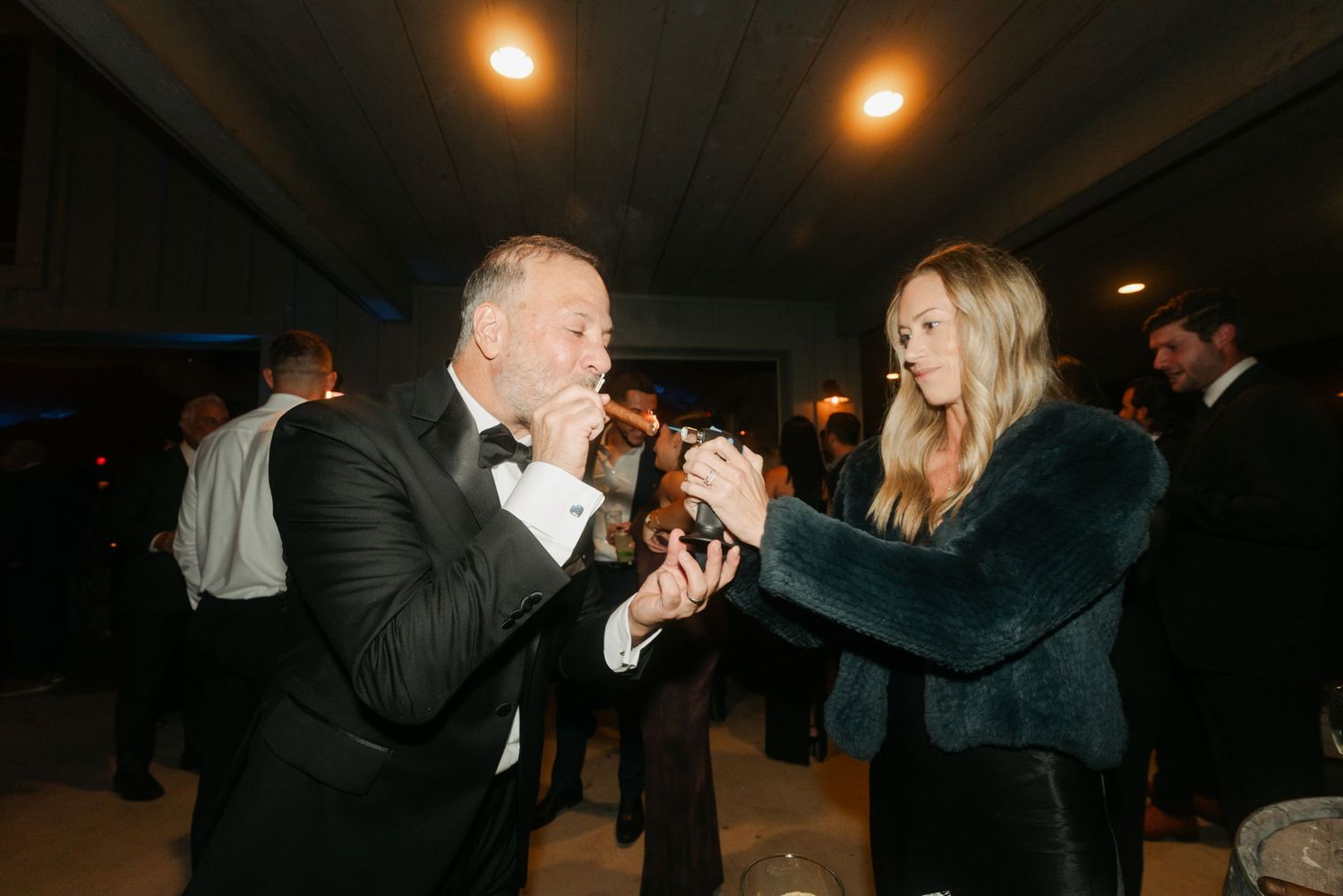 Man in tuxedo kisses woman's hand at elegant evening party, guests socializing in background.