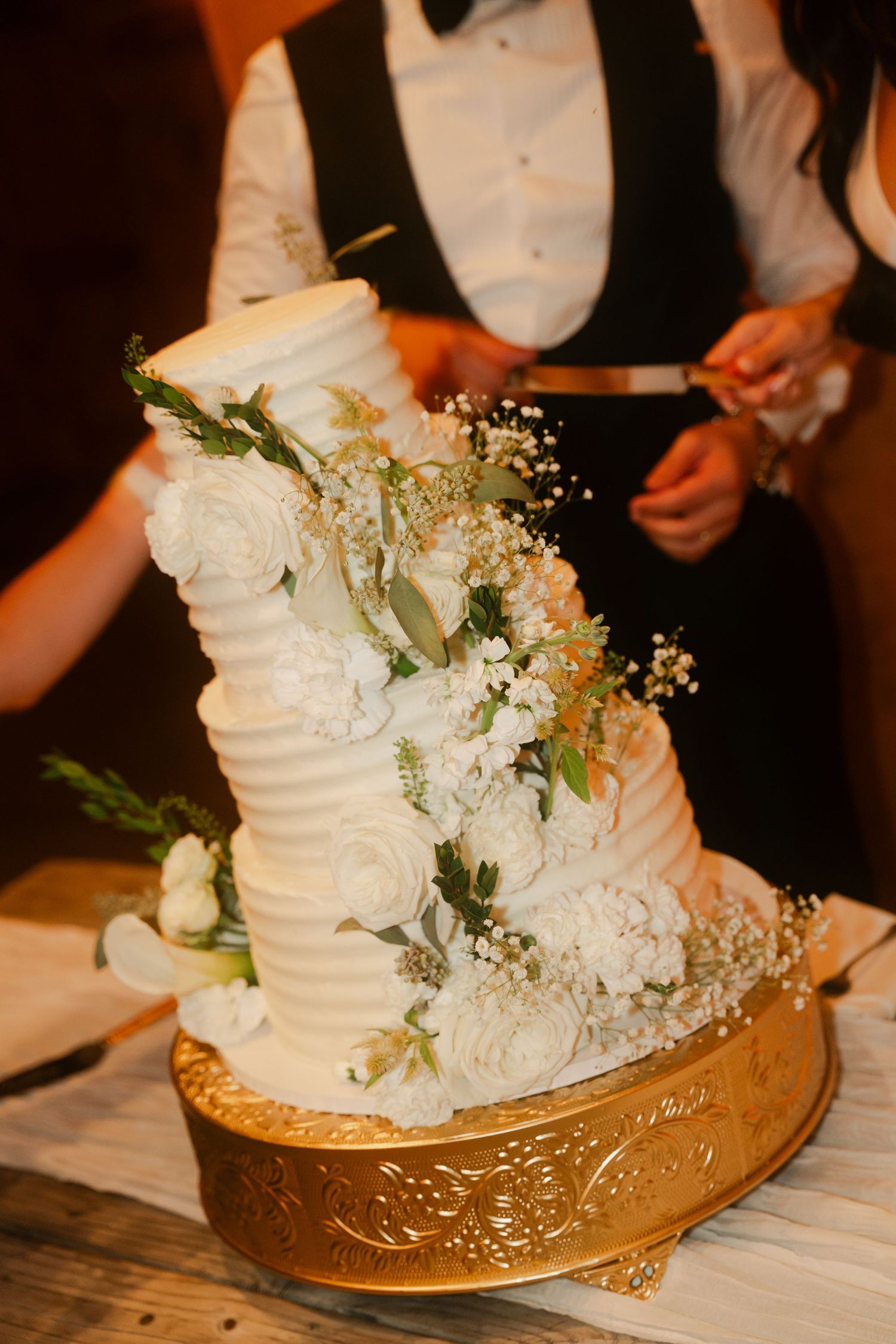 Elegant three-tier white wedding cake adorned with fresh roses and baby's breath on a gold stand.