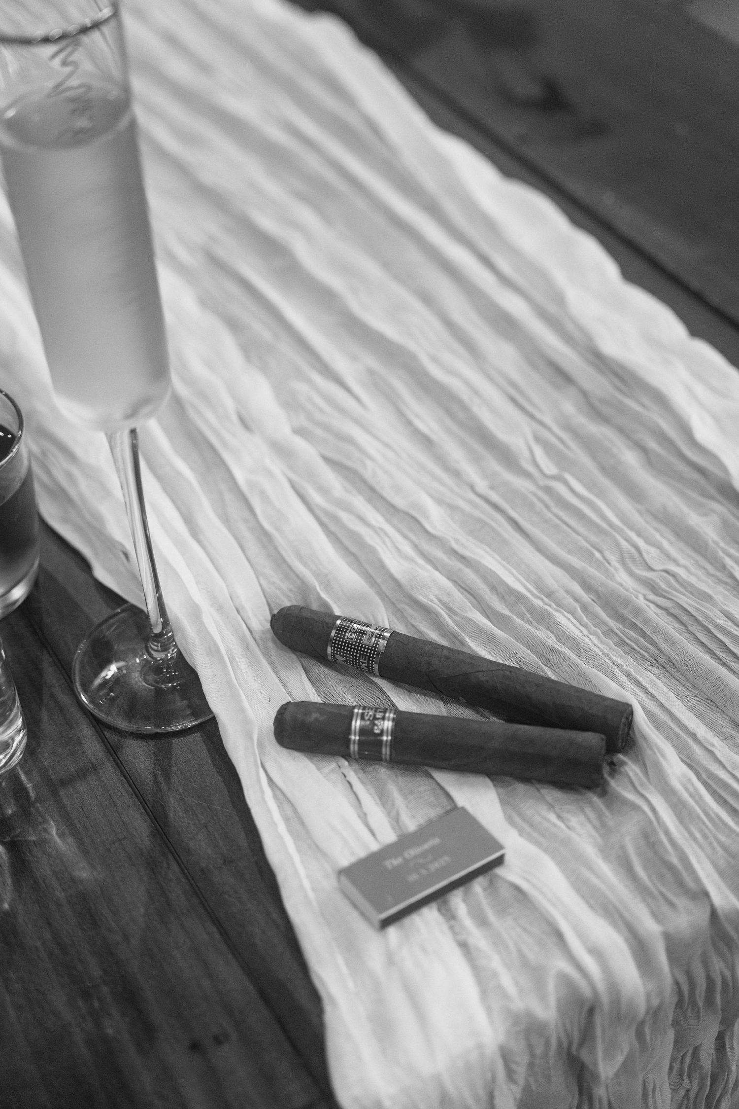 Black and white photo of two cigars and a matchbook on white linen beside champagne glasses.