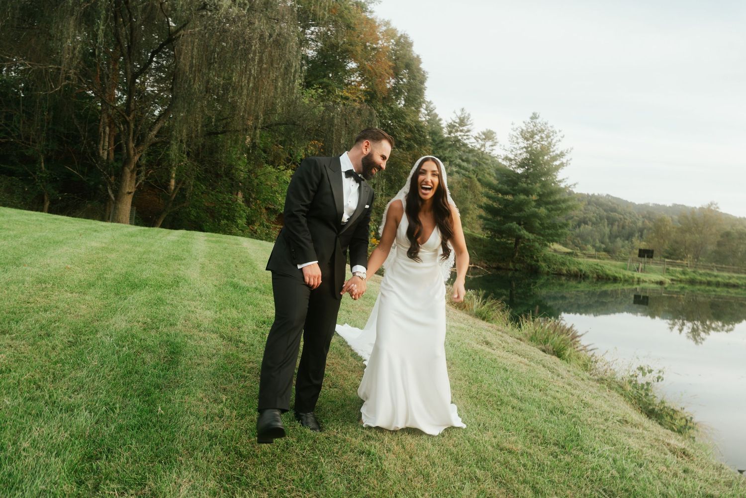 Happy bride and groom walk hand in hand along a lakeside lawn on their wedding day.