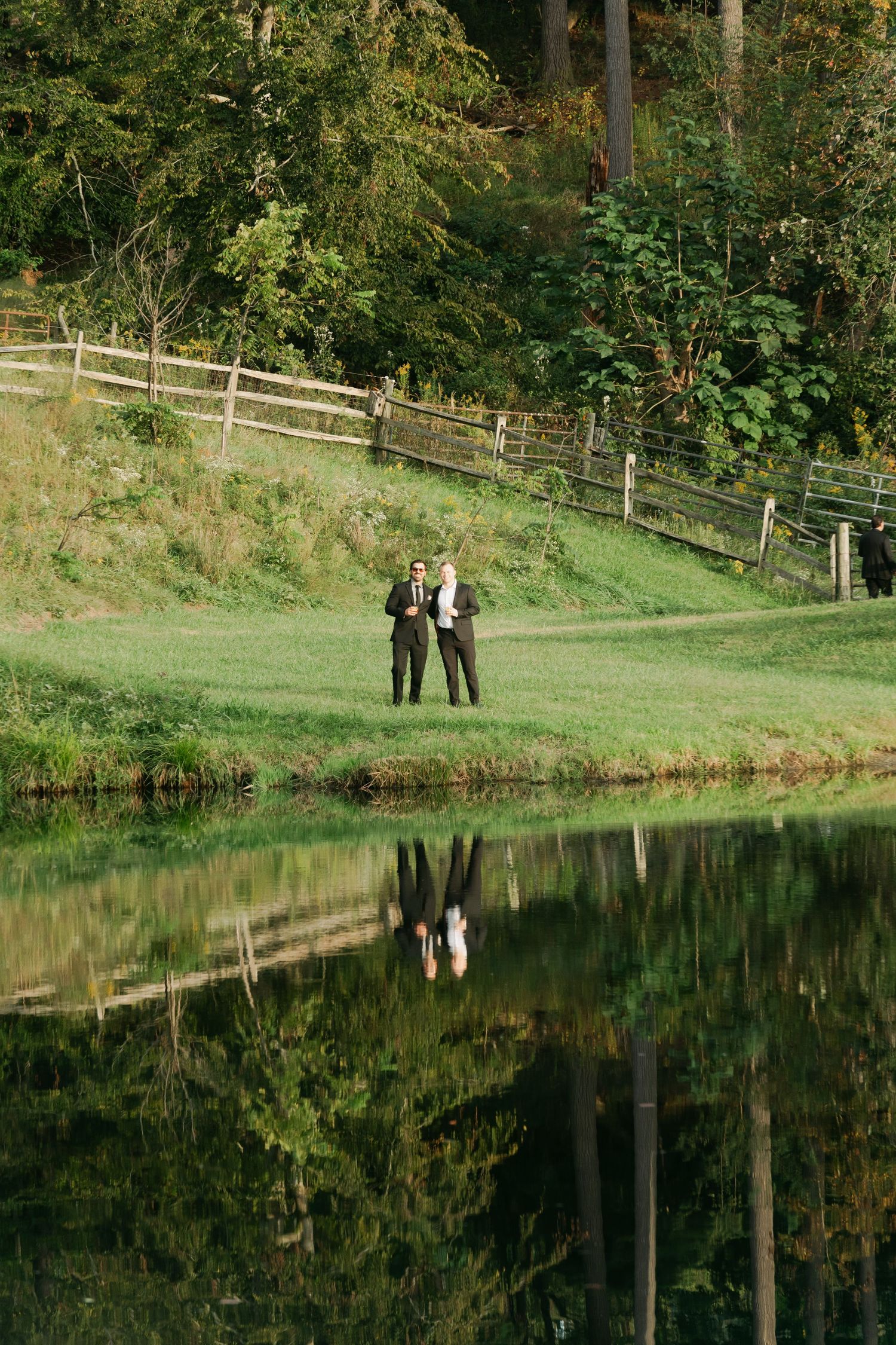 Two people in suits stand by a reflective pond with lush green hills and wooden fence backdrop.