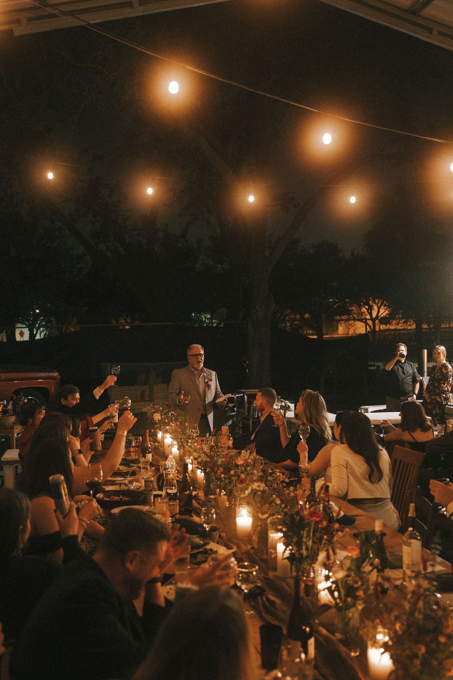 Wedding reception under string lights with guests seated at long tables decorated with floral centerpieces.