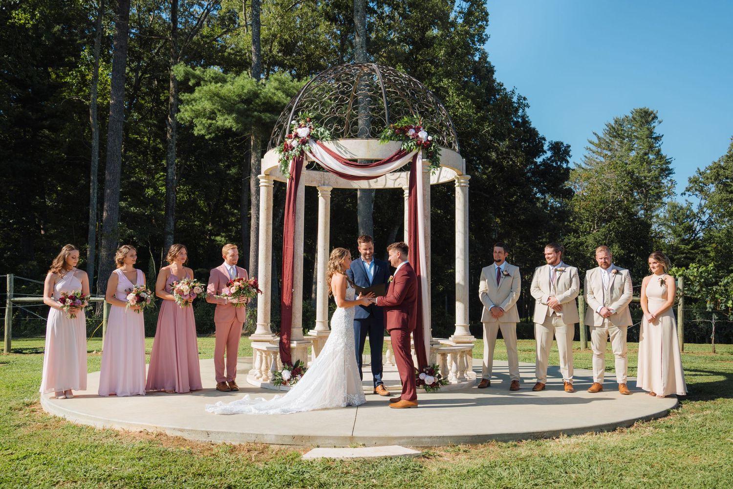 Outdoor wedding ceremony setup with pink bridesmaids and groomsmen lined up under an arch.