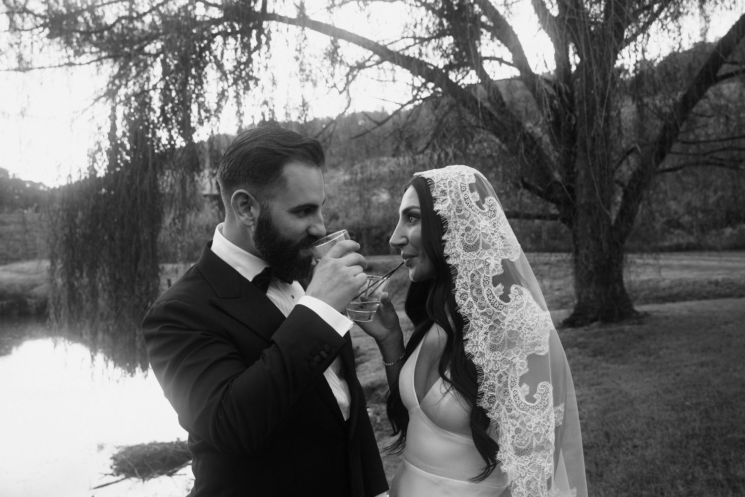 Black and white wedding photo of groom kissing bride's hand near a pond, bride wearing lace veil.