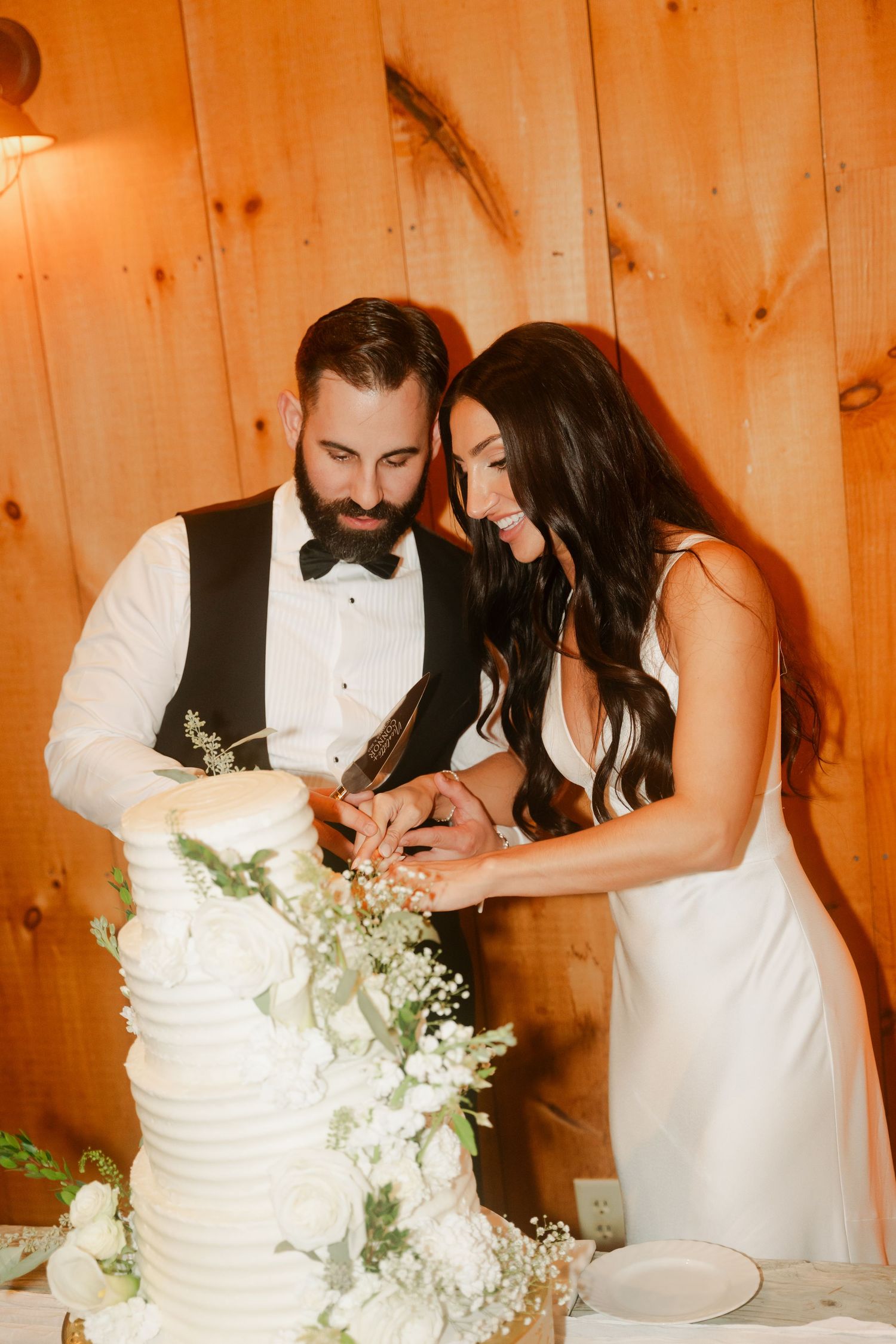 Bride and groom smiling while cutting white tiered wedding cake adorned with greenery.