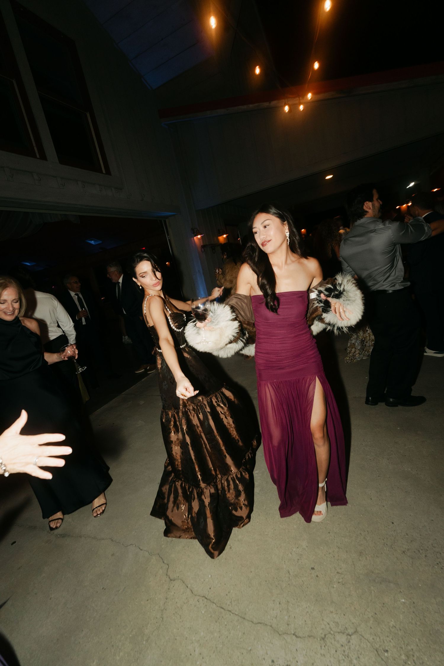 Two elegant women in formal gowns dance with feather accessories at a wedding reception.