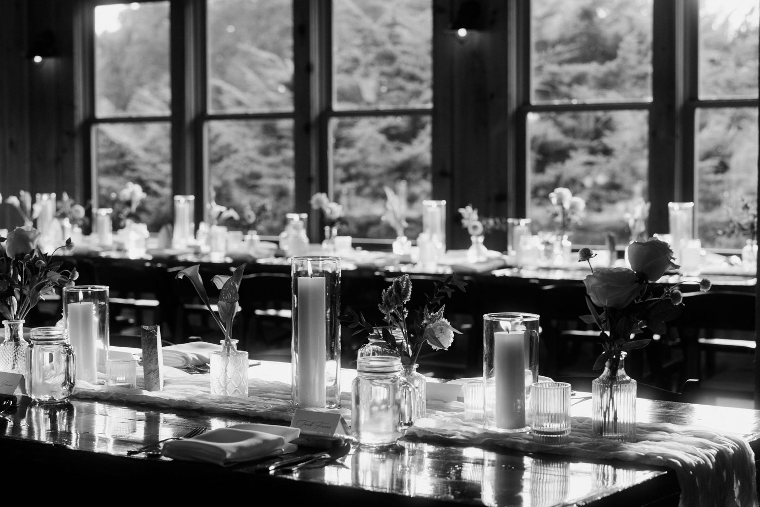 Black and white photo of elegant wedding reception table with candles and floral arrangements near large windows.