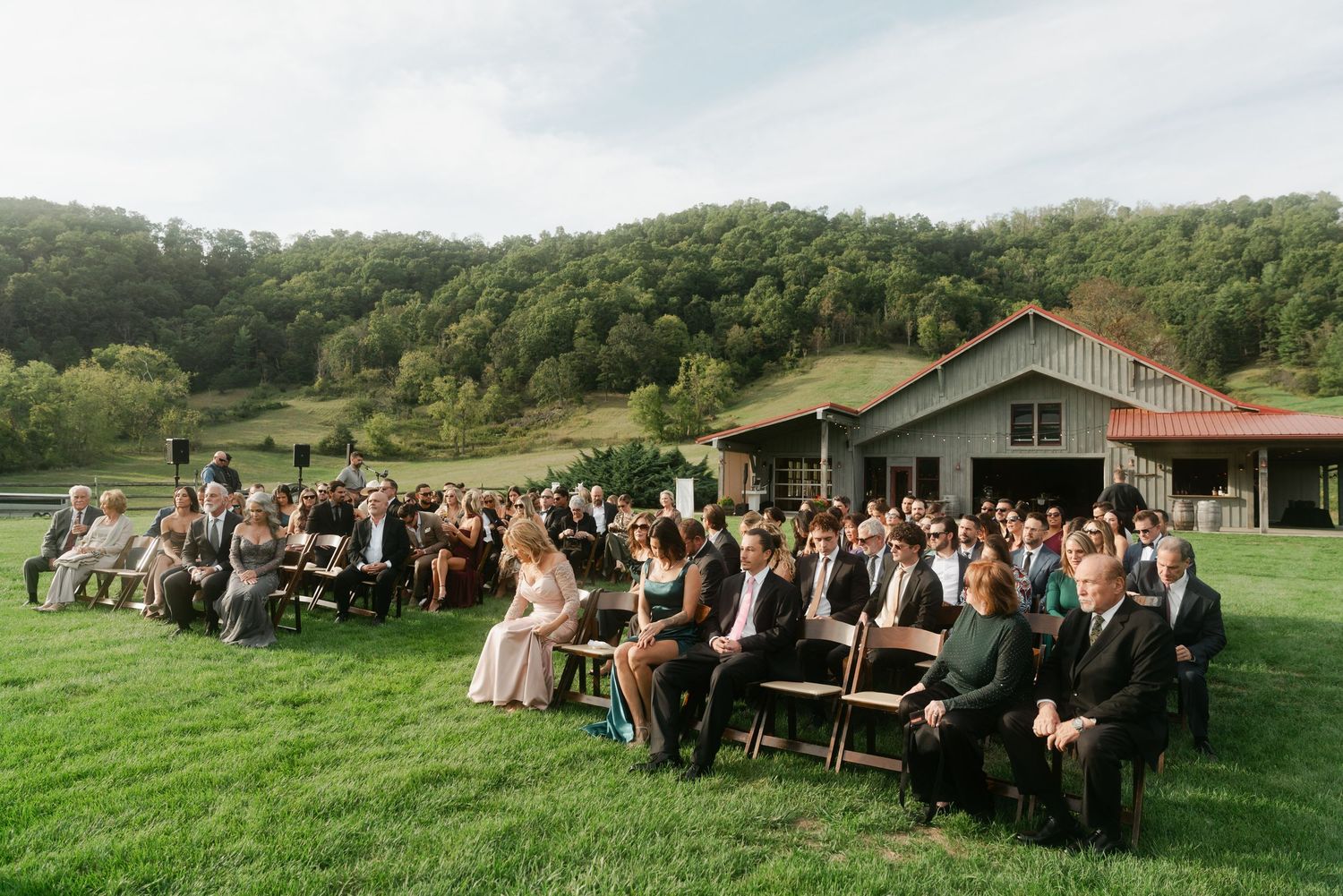Outdoor wedding ceremony with guests seated on wooden chairs facing a rustic barn amid rolling hills.