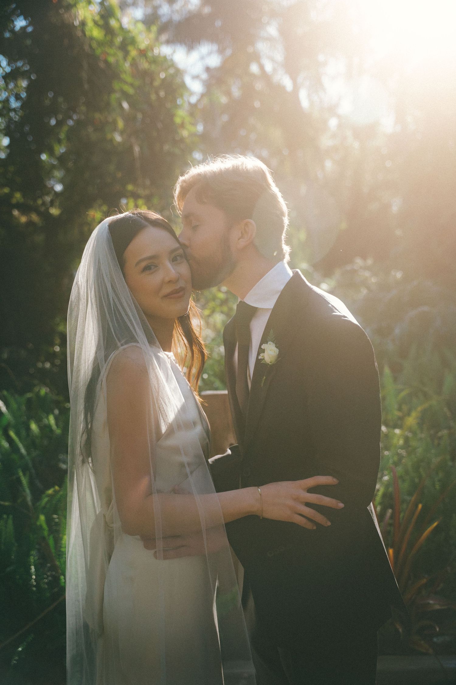 Romantic wedding couple embracing in a sun-drenched garden setting during golden hour.