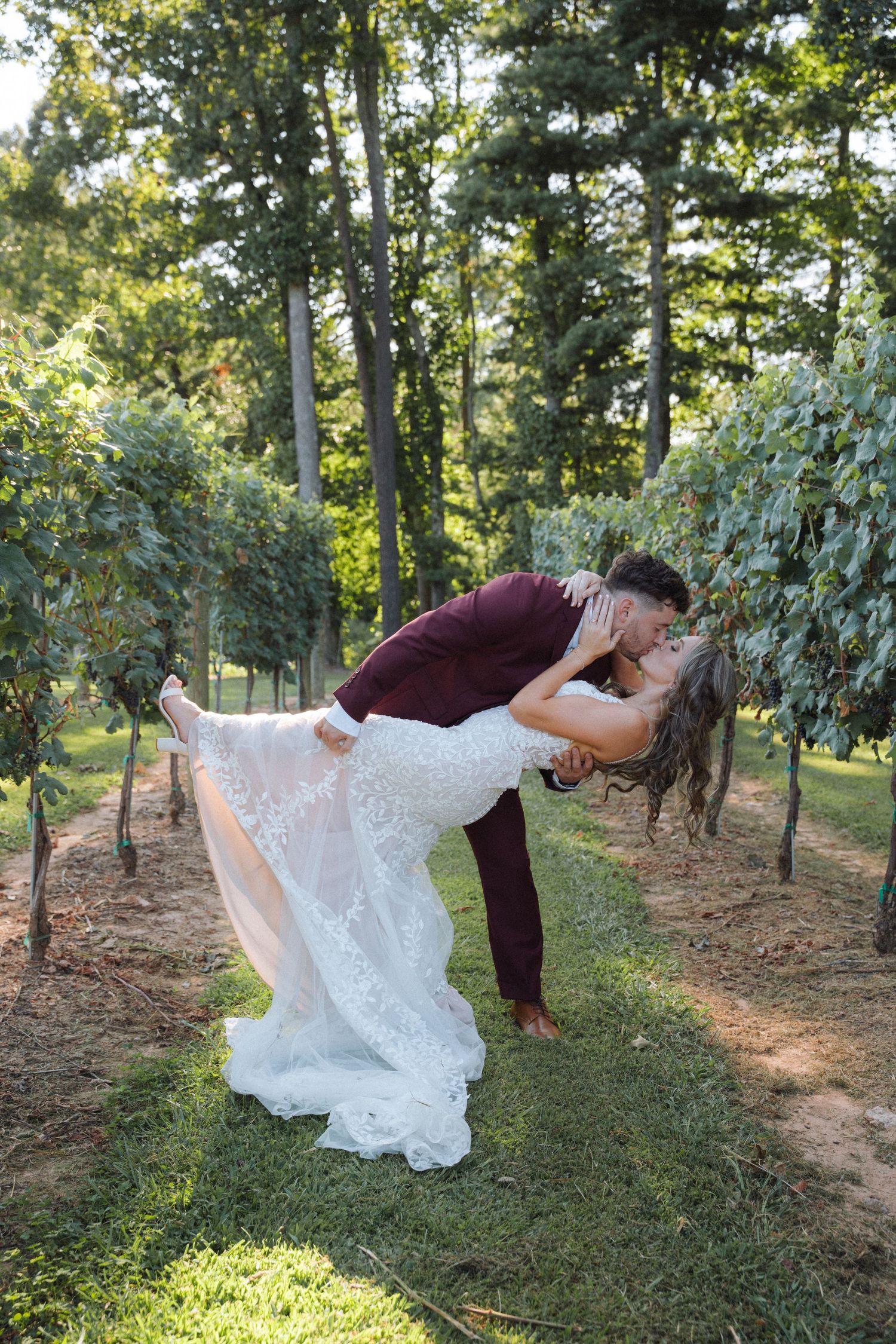 A couple performs a dramatic dip pose in their wedding attire among sunlit trees and greenery.