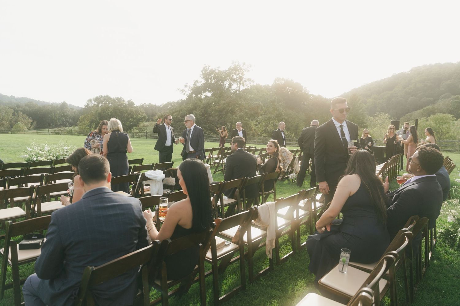 Outdoor wedding ceremony with guests seated on wooden chairs on a green lawn surrounded by rolling hills.