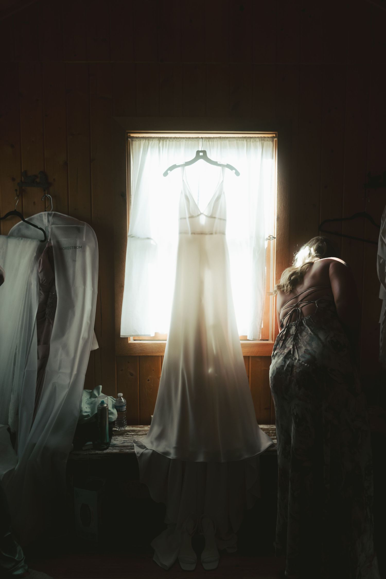 Elegant wedding dress hanging in window light with bridal party getting ready in rustic wooden room.