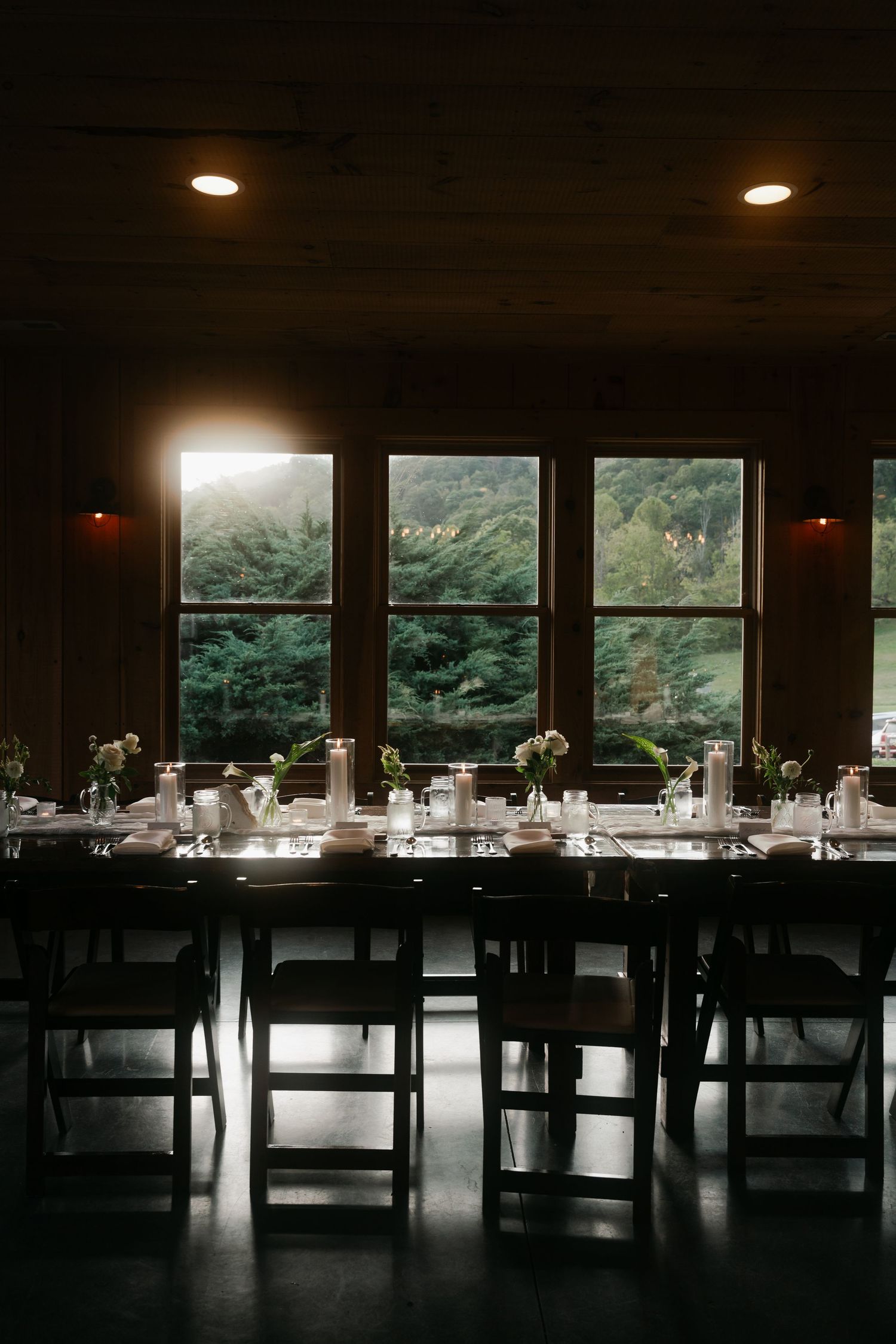 Dimly lit rustic wedding reception hall with long tables, folding chairs, and forest views.