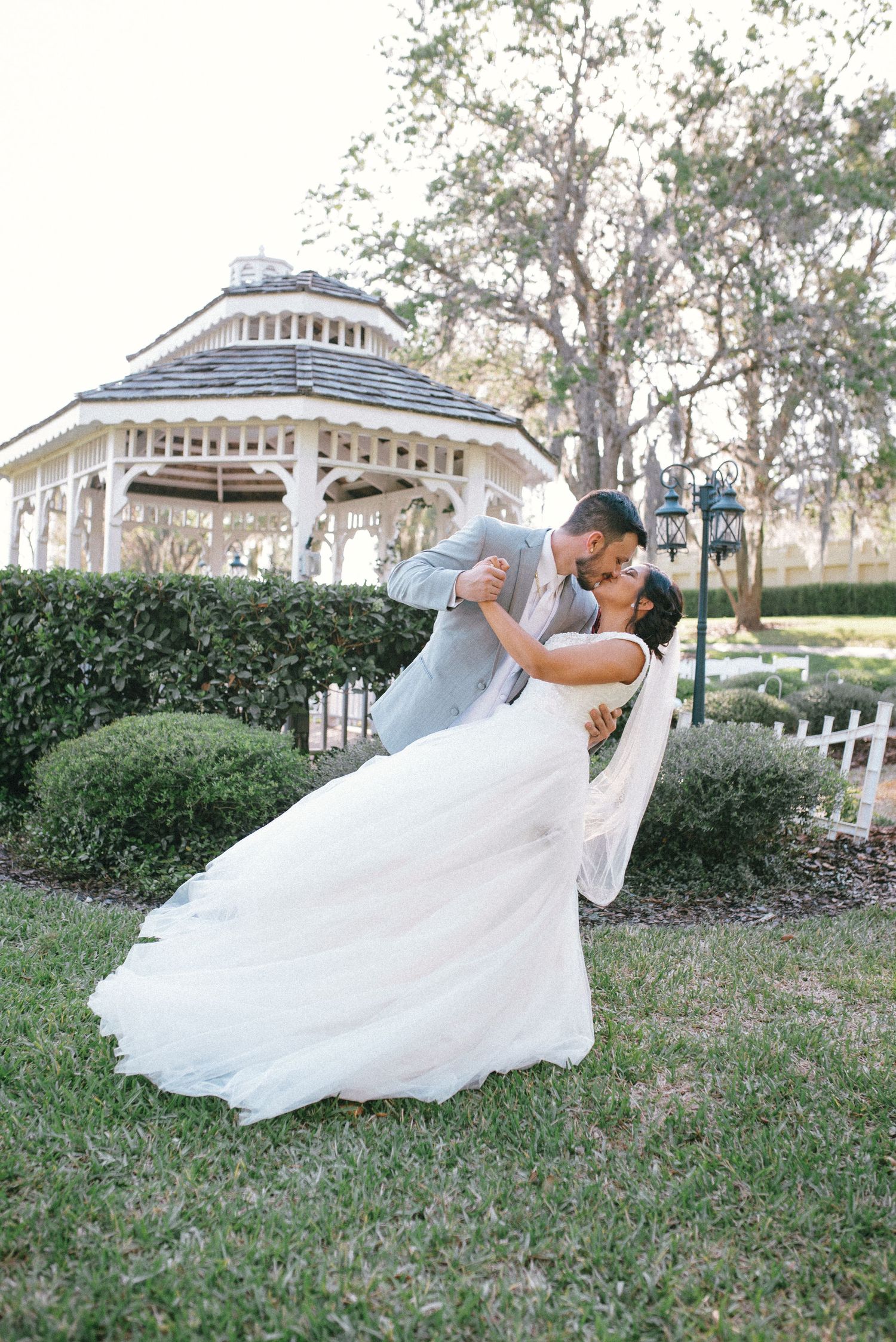 Romantic wedding dip pose in front of a white gazebo surrounded by landscaped gardens.
