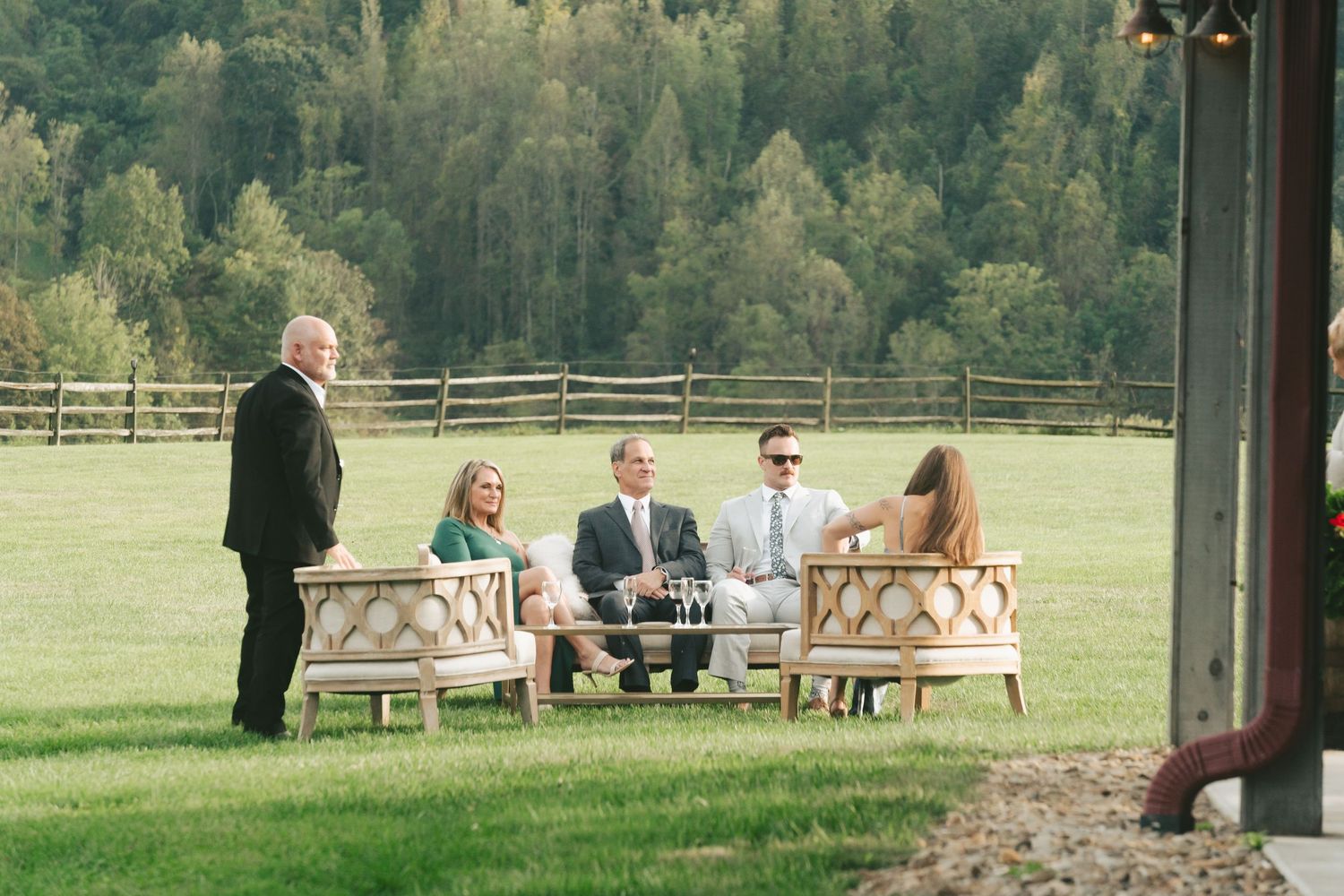Wedding guests relaxing on outdoor lounge furniture on green lawn at rustic farm venue.