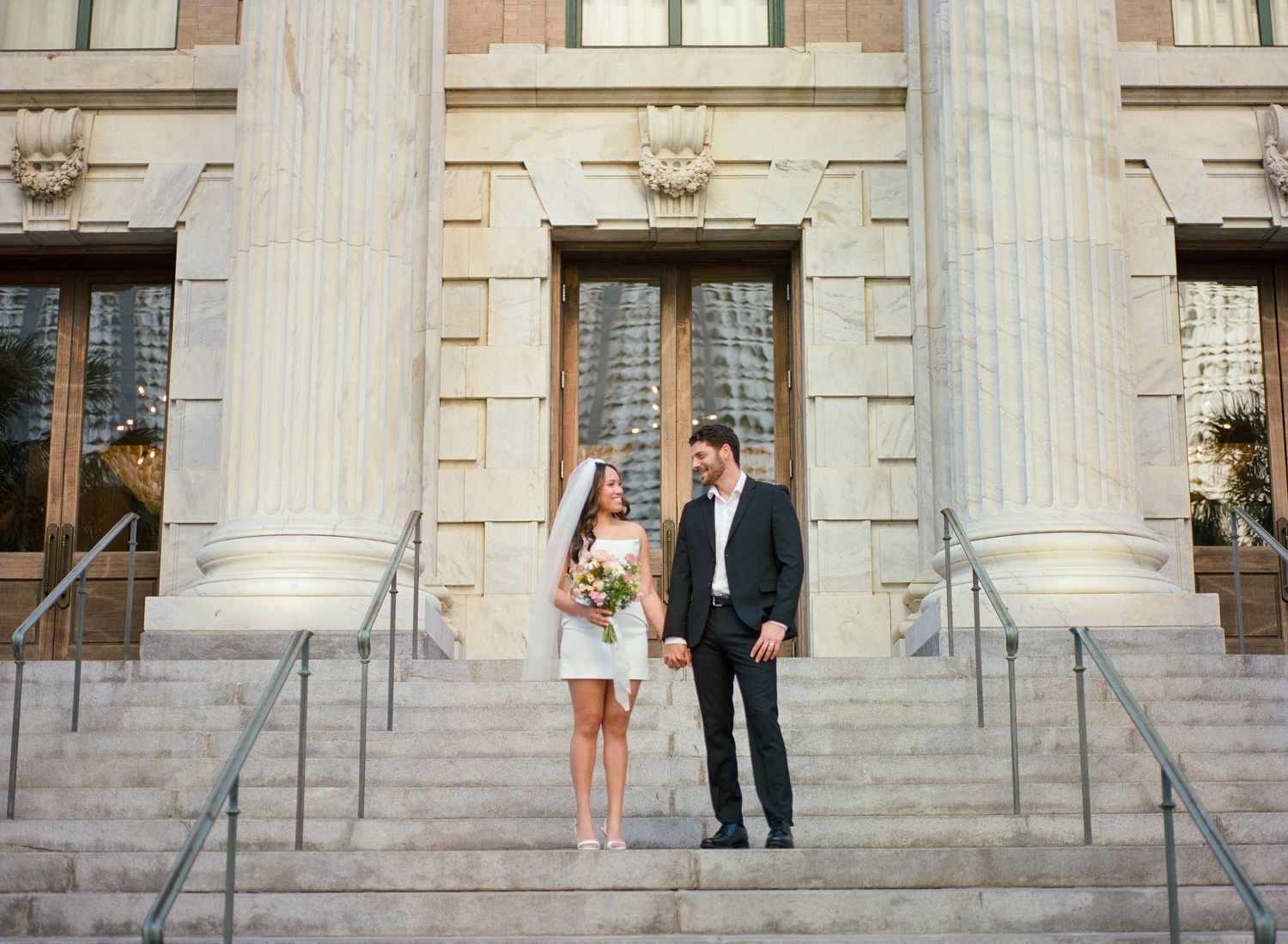 Bride and groom stand on steps of grand columned building, bride in short white dress holding colorful bouquet.