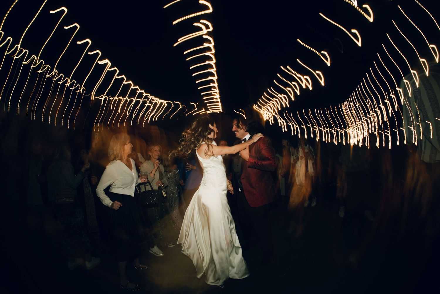 Bride and groom share first dance surrounded by swirling light trails at a dark romantic wedding reception.