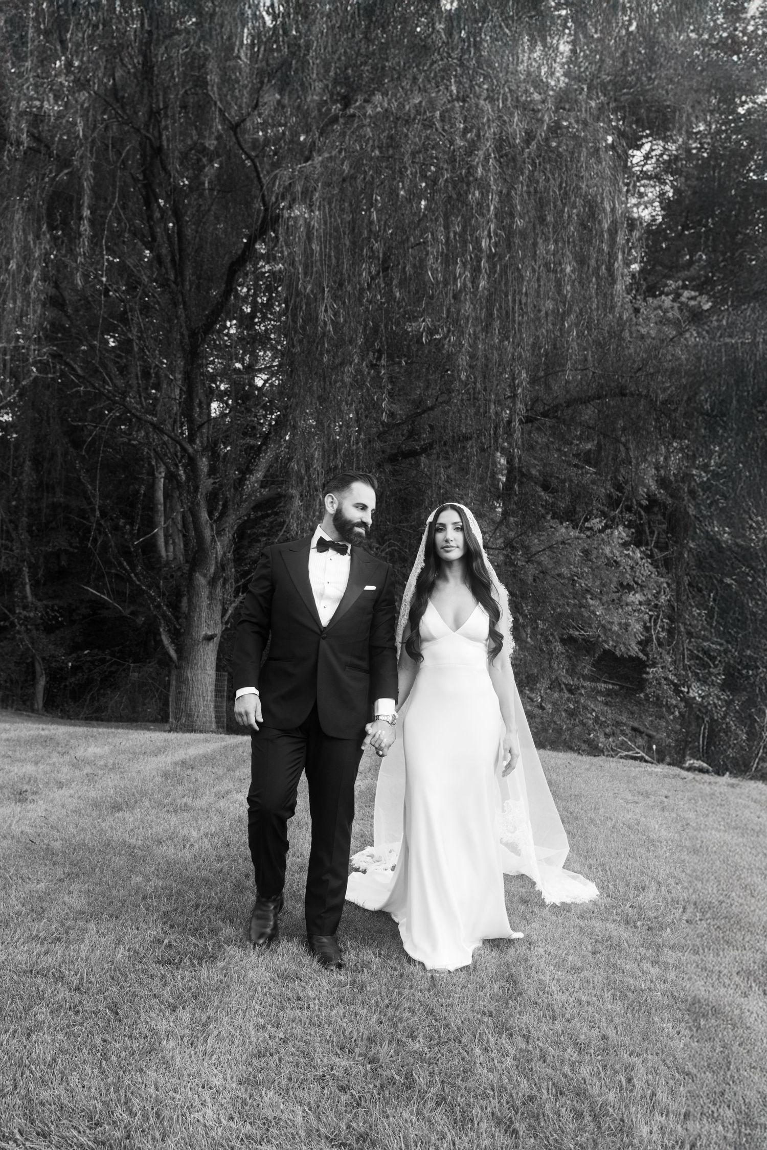 Black and white photo of bride and groom holding hands on a grassy lawn with willow trees.