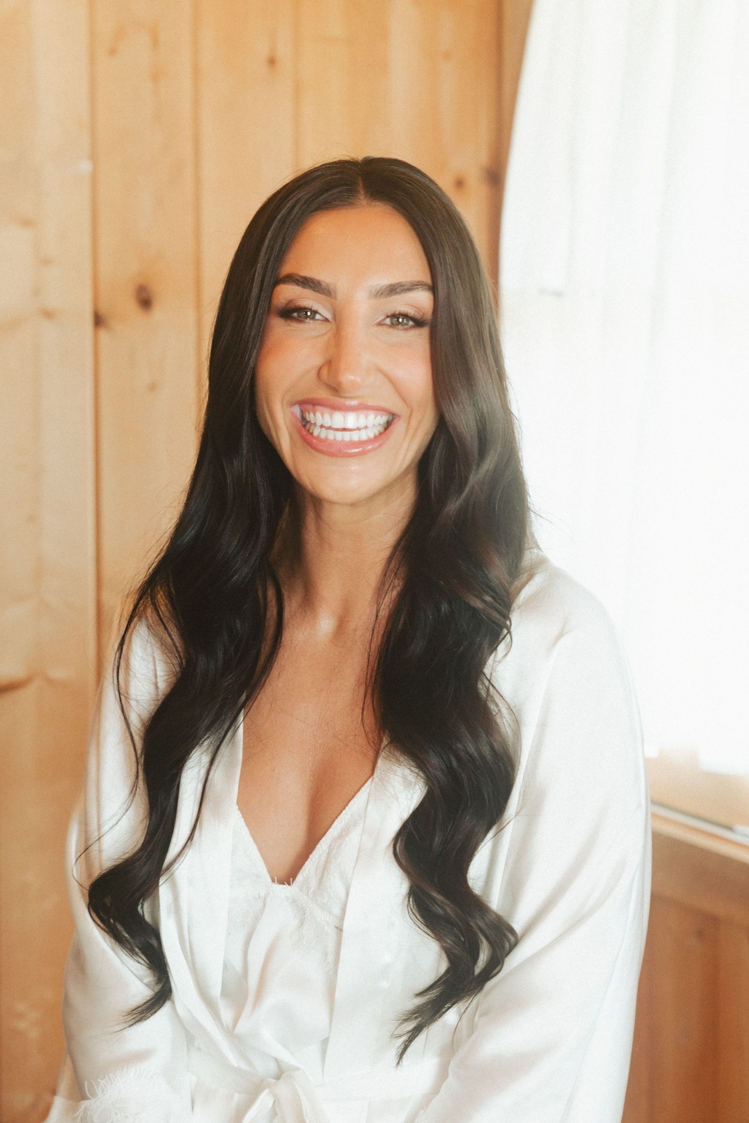 A smiling bride with long dark wavy hair wearing a white satin robe in a warmly lit wooden room.