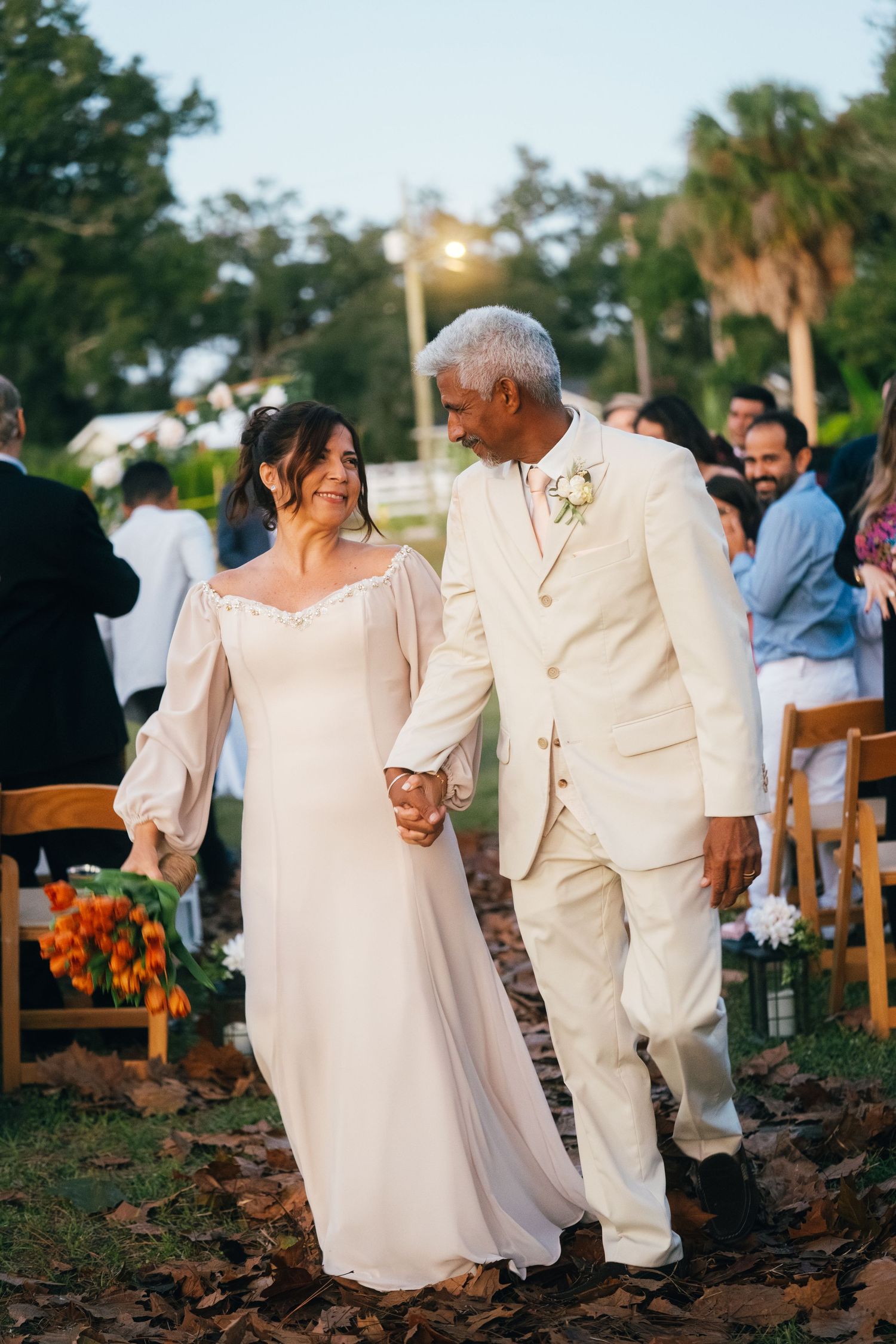 An elderly couple in white outfits walking together at an outdoor wedding celebration.