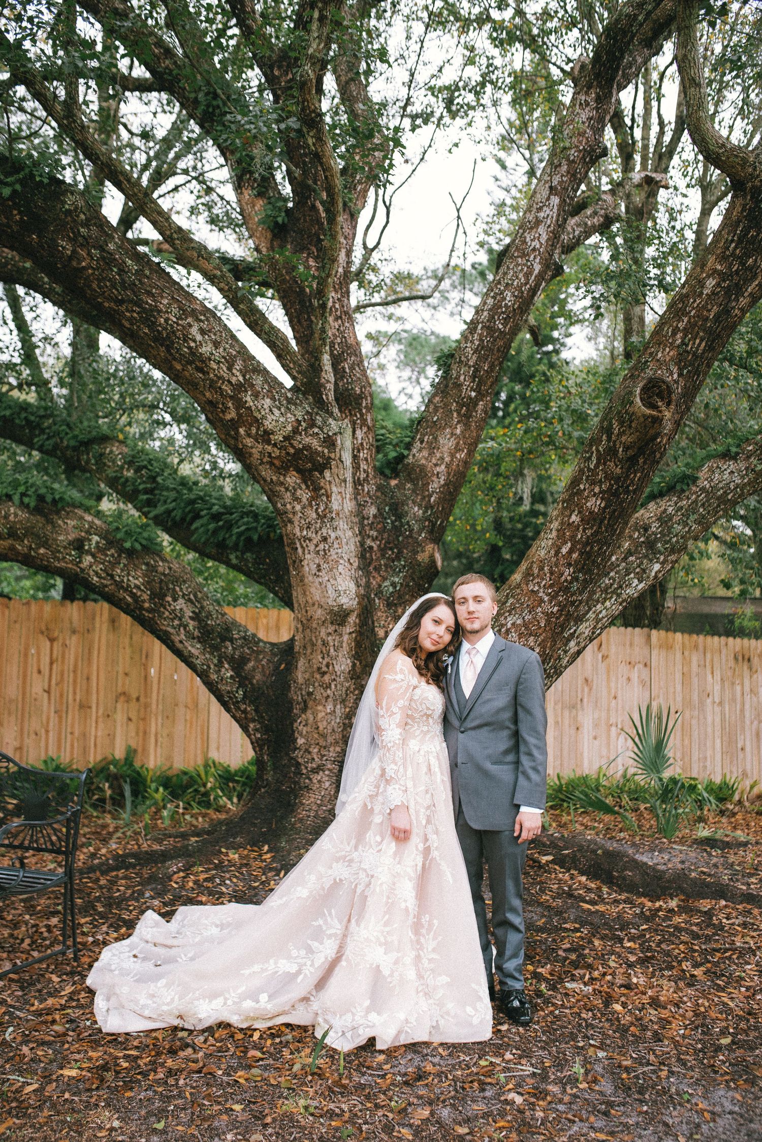 A couple poses for their wedding portrait in front of a large branching tree in a garden setting.