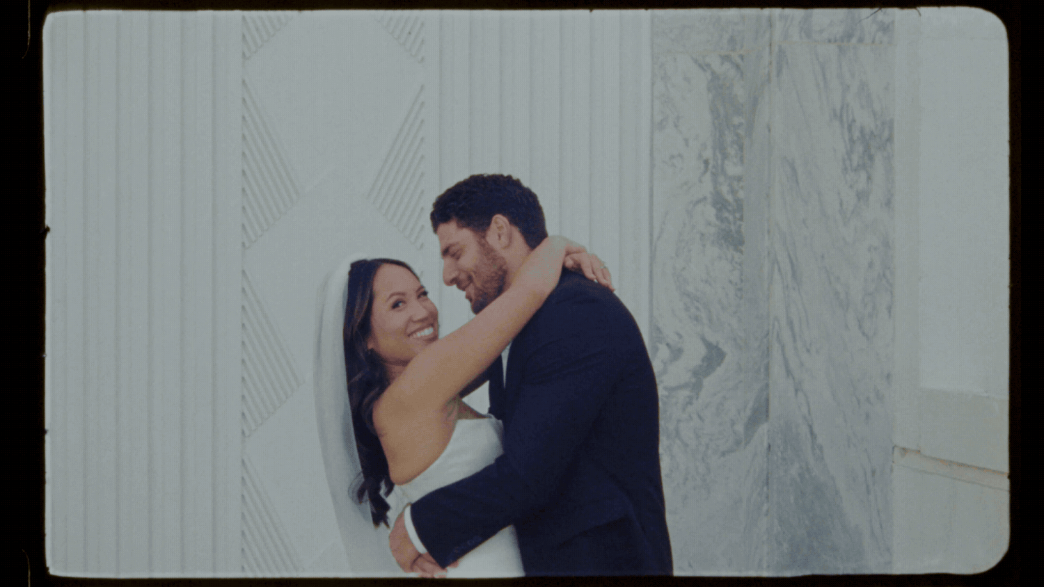 Happy couple embracing and smiling on their wedding day, bride in white gown and groom in navy suit.