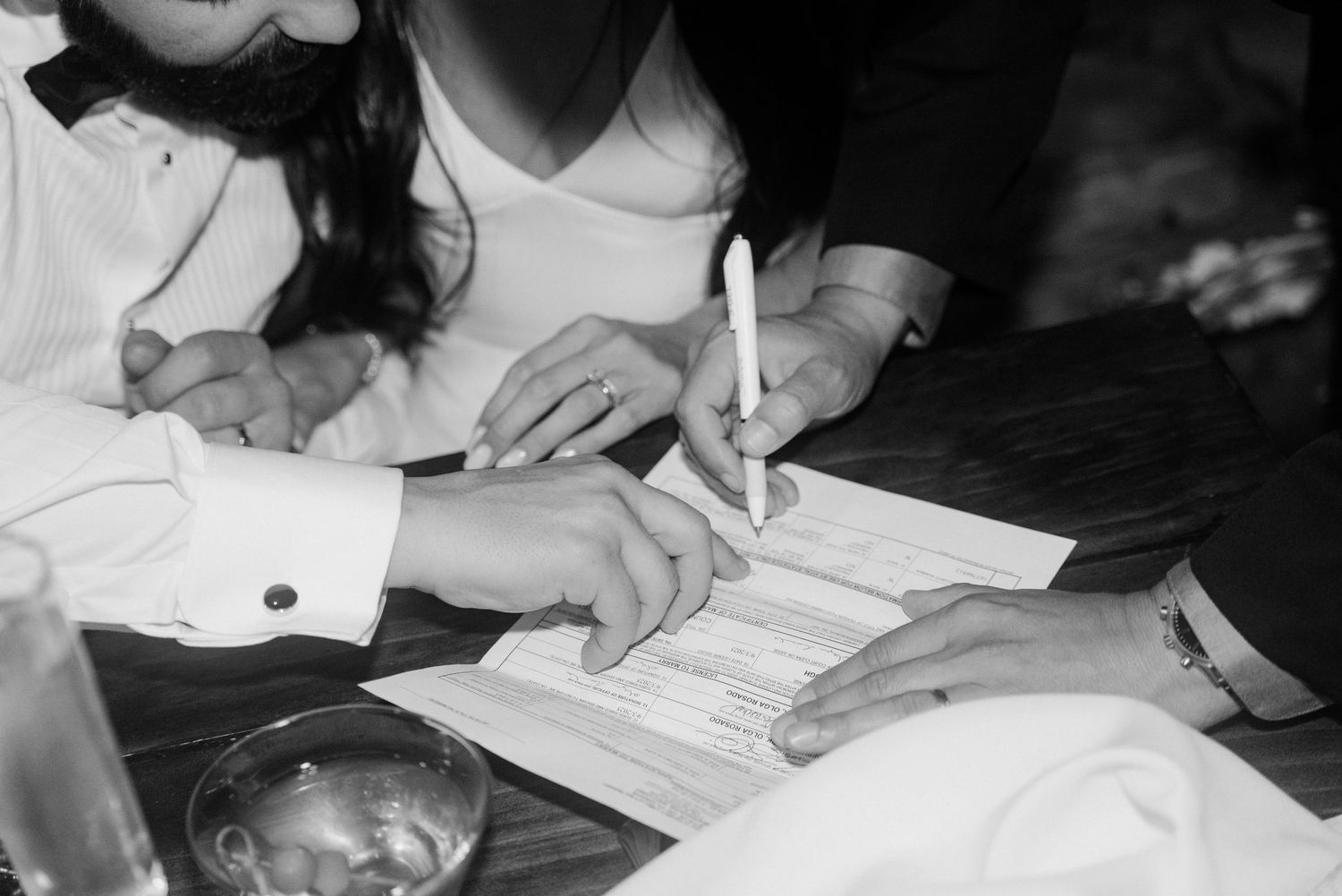Black and white close-up of bride and groom signing wedding certificate with pen at reception.