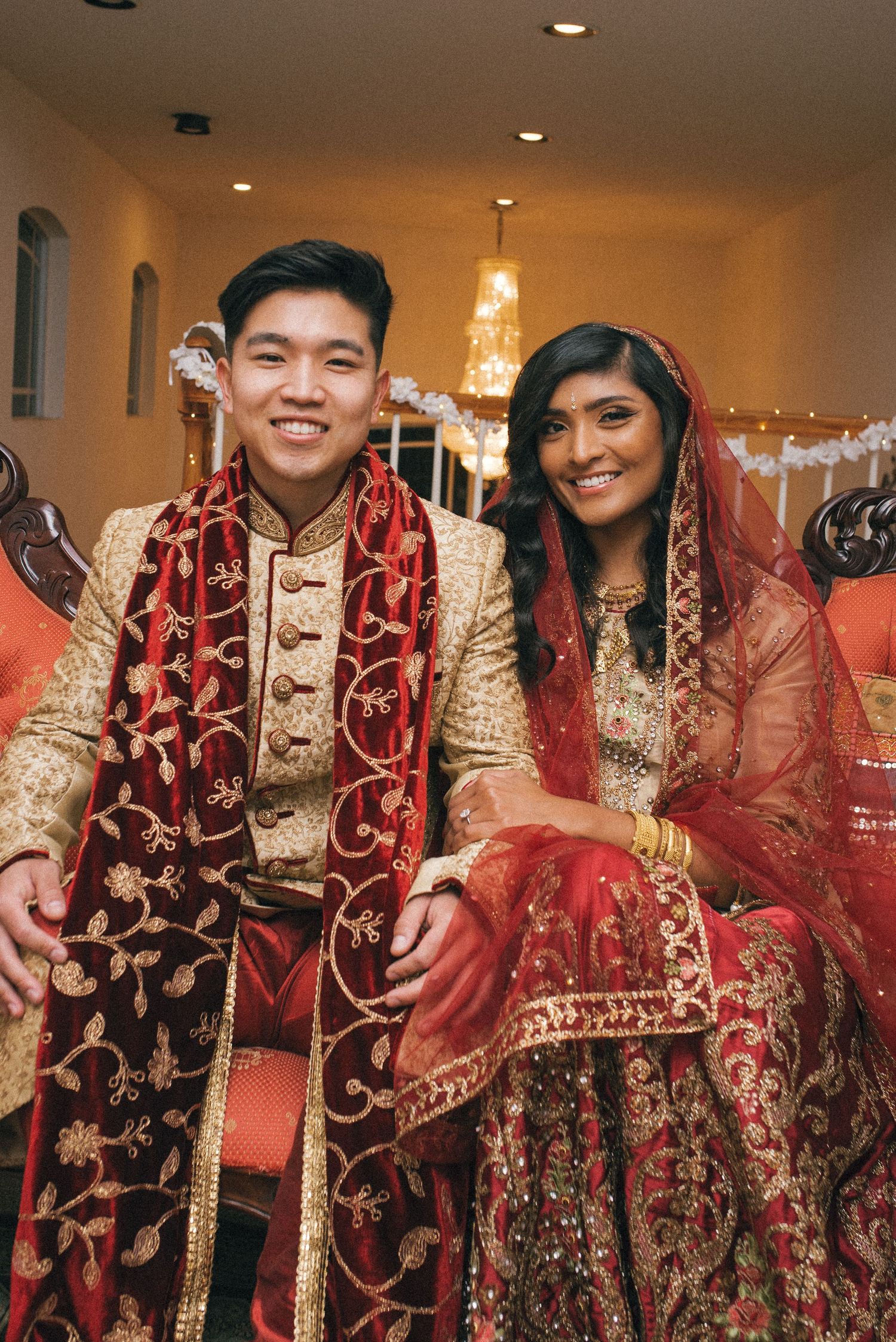 A couple wearing traditional South Asian wedding attire pose together indoors with decorative lighting.