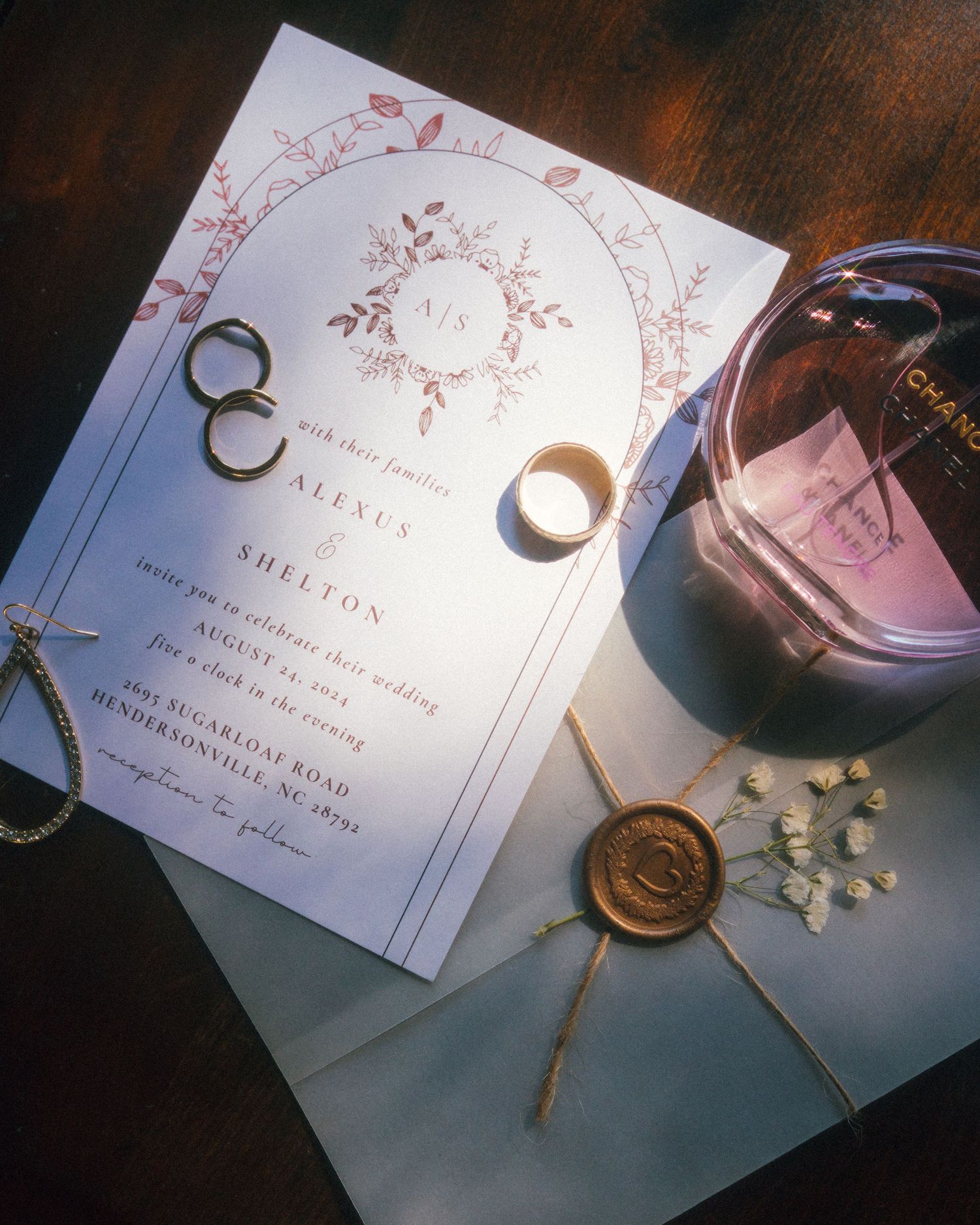 Wedding invitation with floral design, rings, wax seal and perfume bottle on dark surface.
