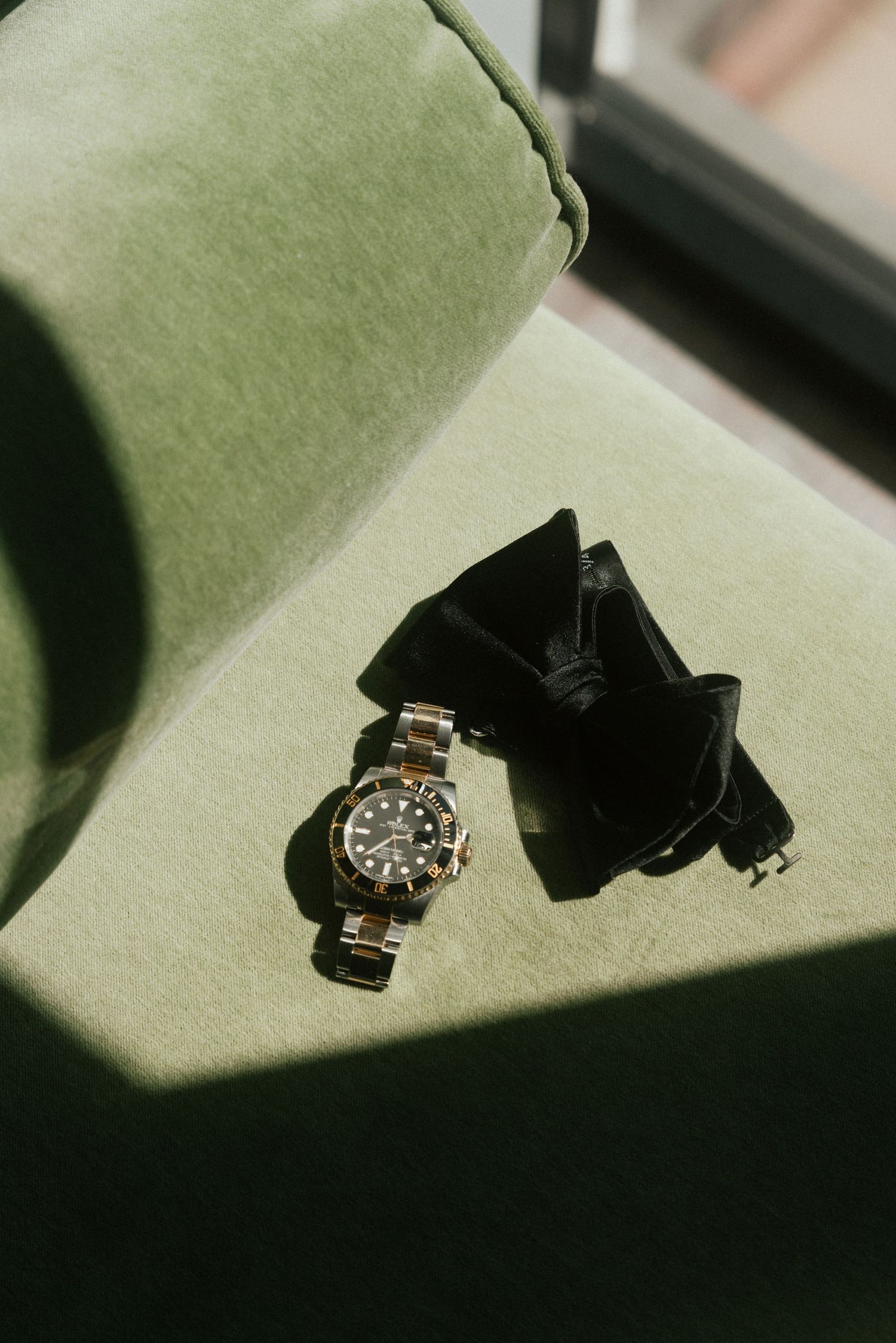 Rolex watch with black dial and two-tone bracelet resting on green velvet surface beside black bow tie.