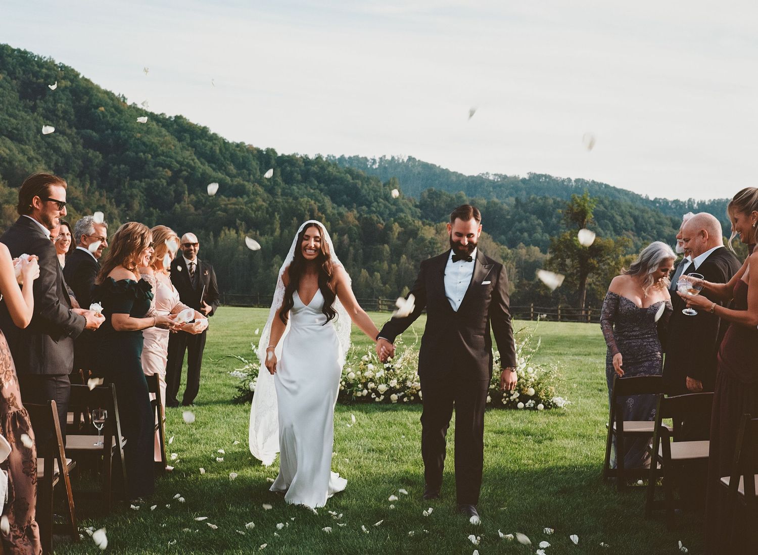 Outdoor wedding celebration in a green mountainside setting with guests gathered around.