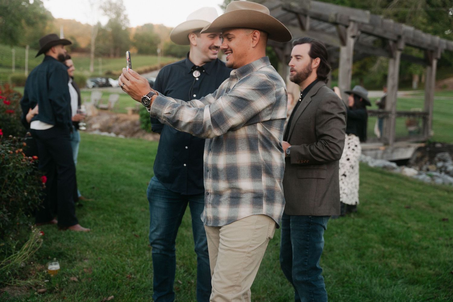 Men in cowboy hats taking a selfie outdoors at a rustic countryside gathering during golden hour.