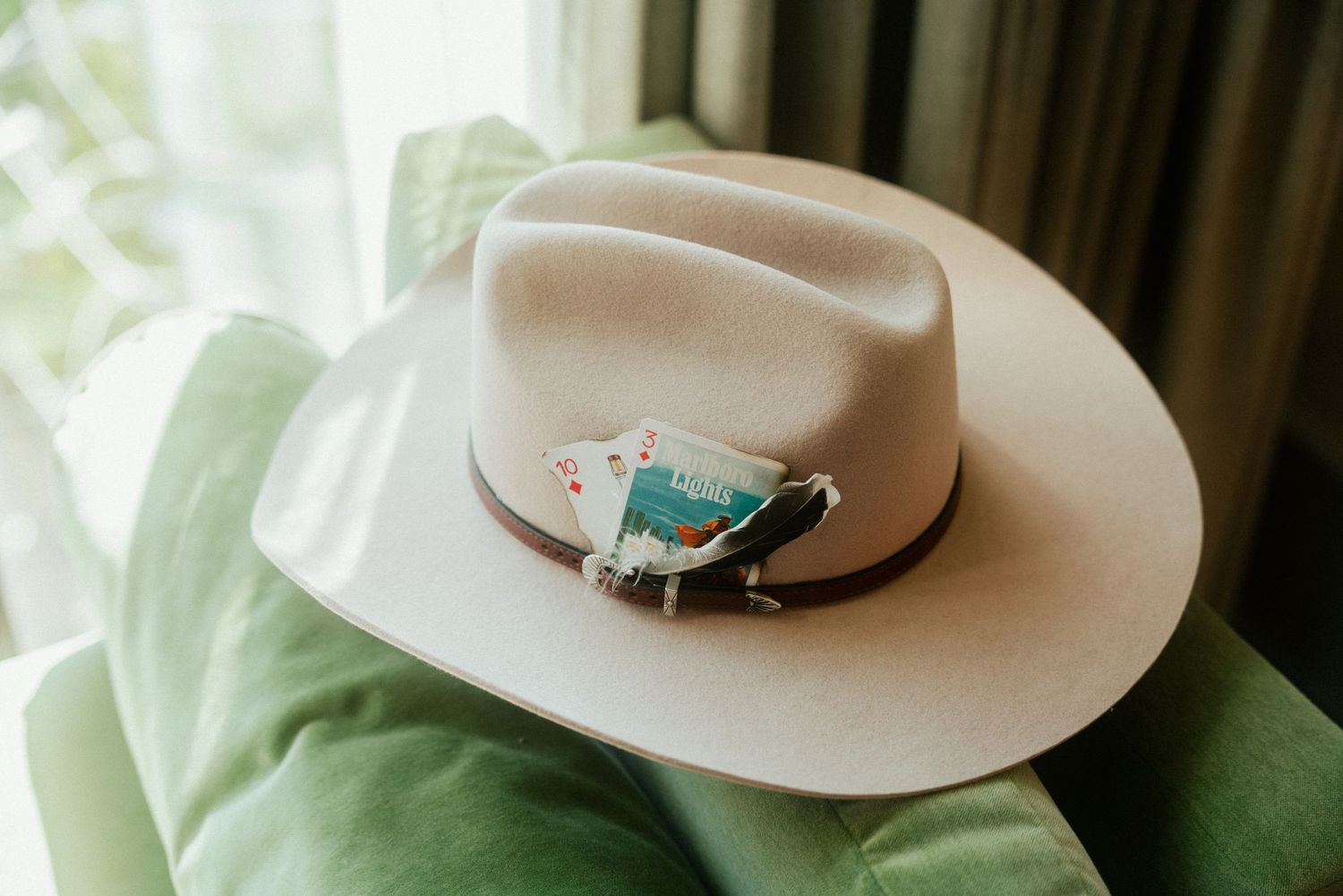 A cream felt cowboy hat with a decorative feather pin resting on a green velvet chair near a window.