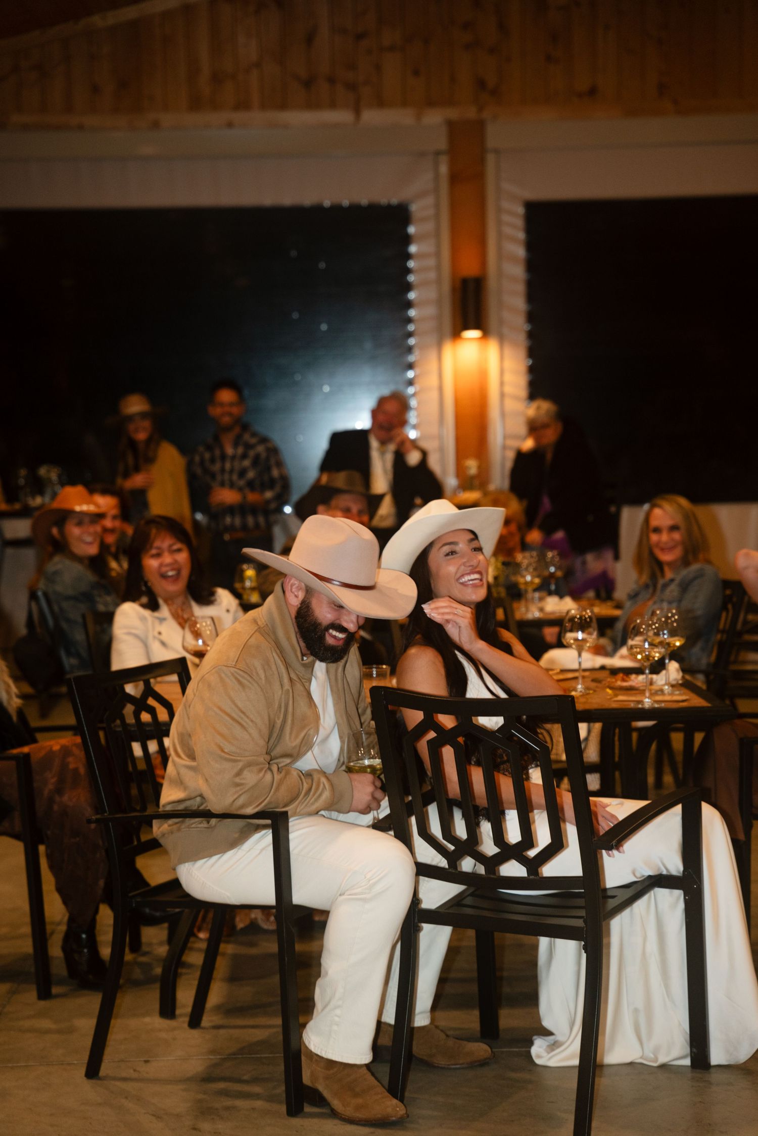 Couple in white cowboy hats laughing at a rustic wedding reception dinner with guests in background.