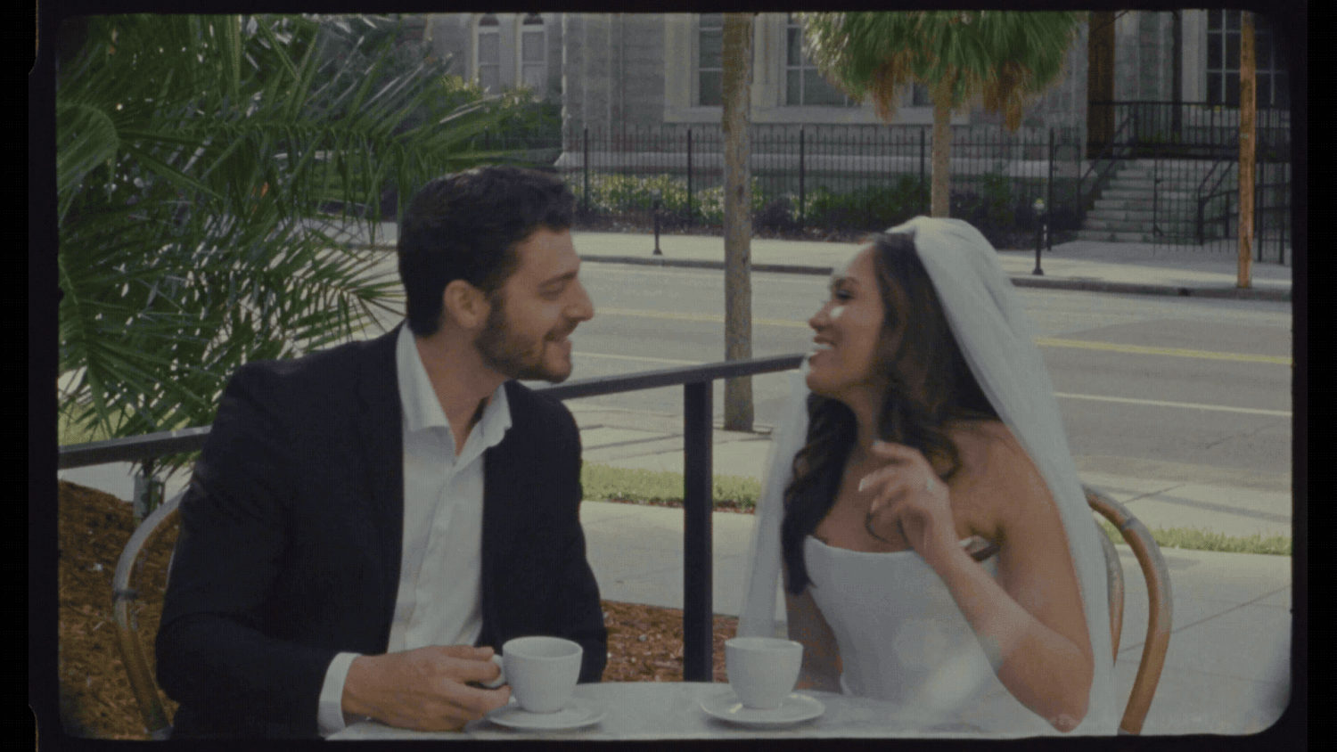 A bride and groom share a romantic moment at an outdoor cafe table, smiling at each other over coffee cups.