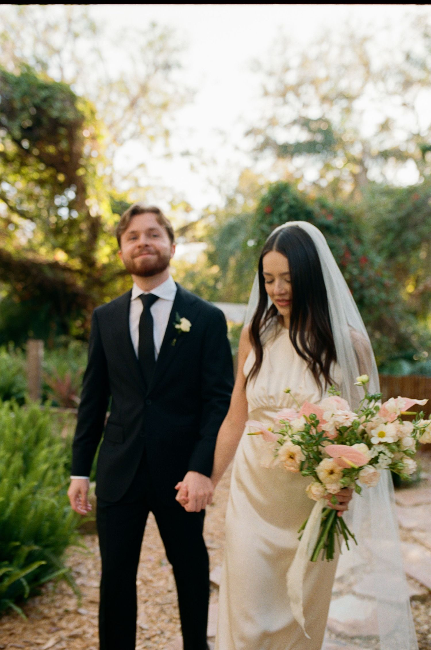 Newlywed couple walking hand in hand through garden with bridal bouquet and formal attire.