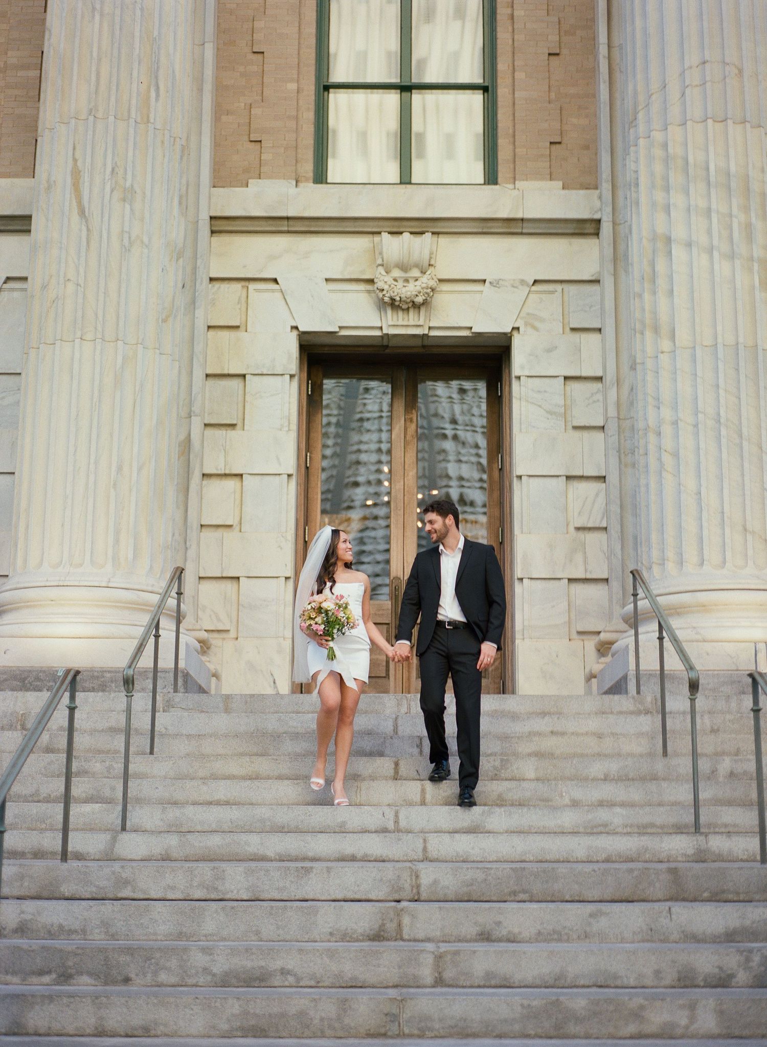 A couple poses on the grand steps of a neoclassical building with white columns.