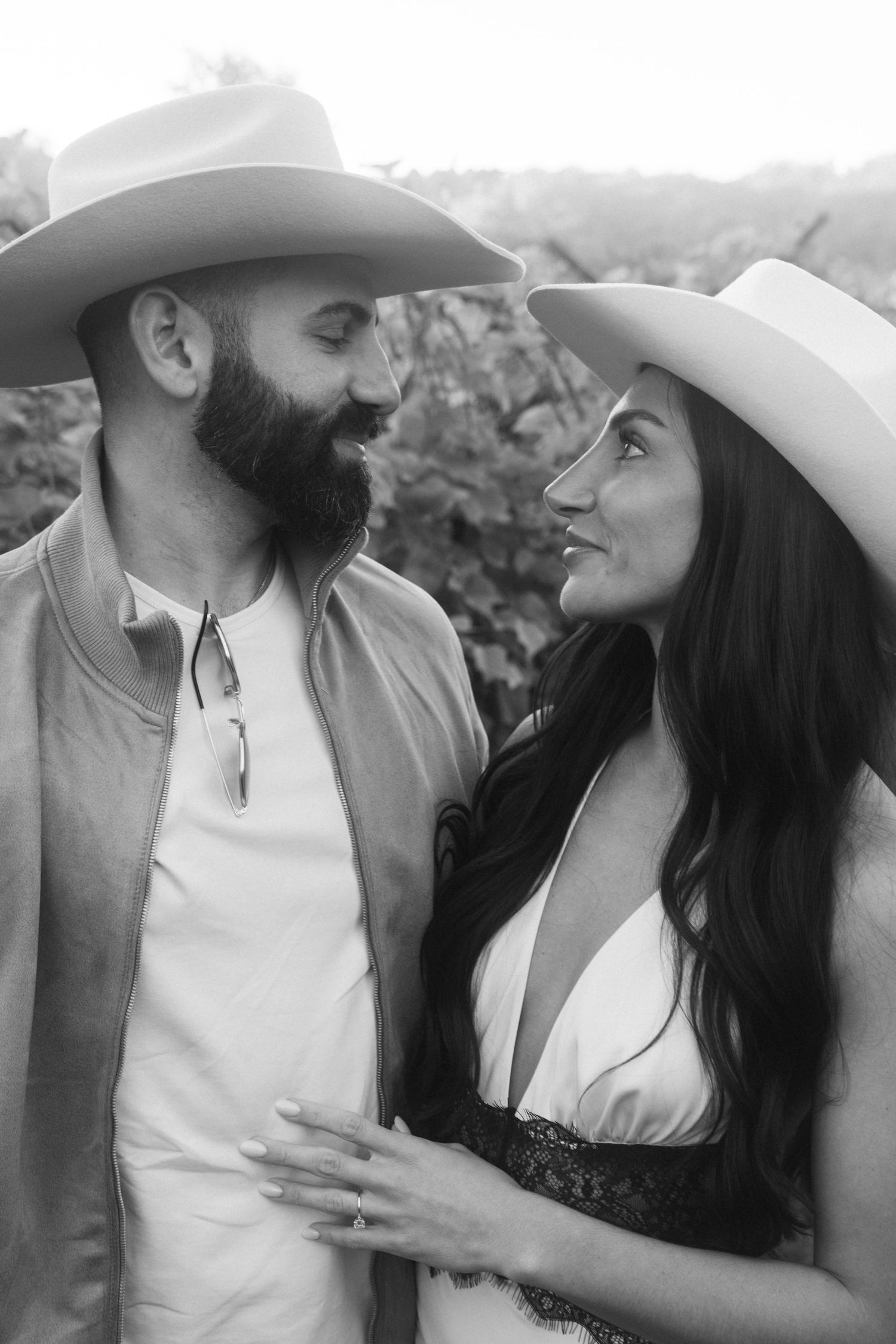 Black and white photo of a bearded man and woman in cowboy hats smiling at each other outdoors.