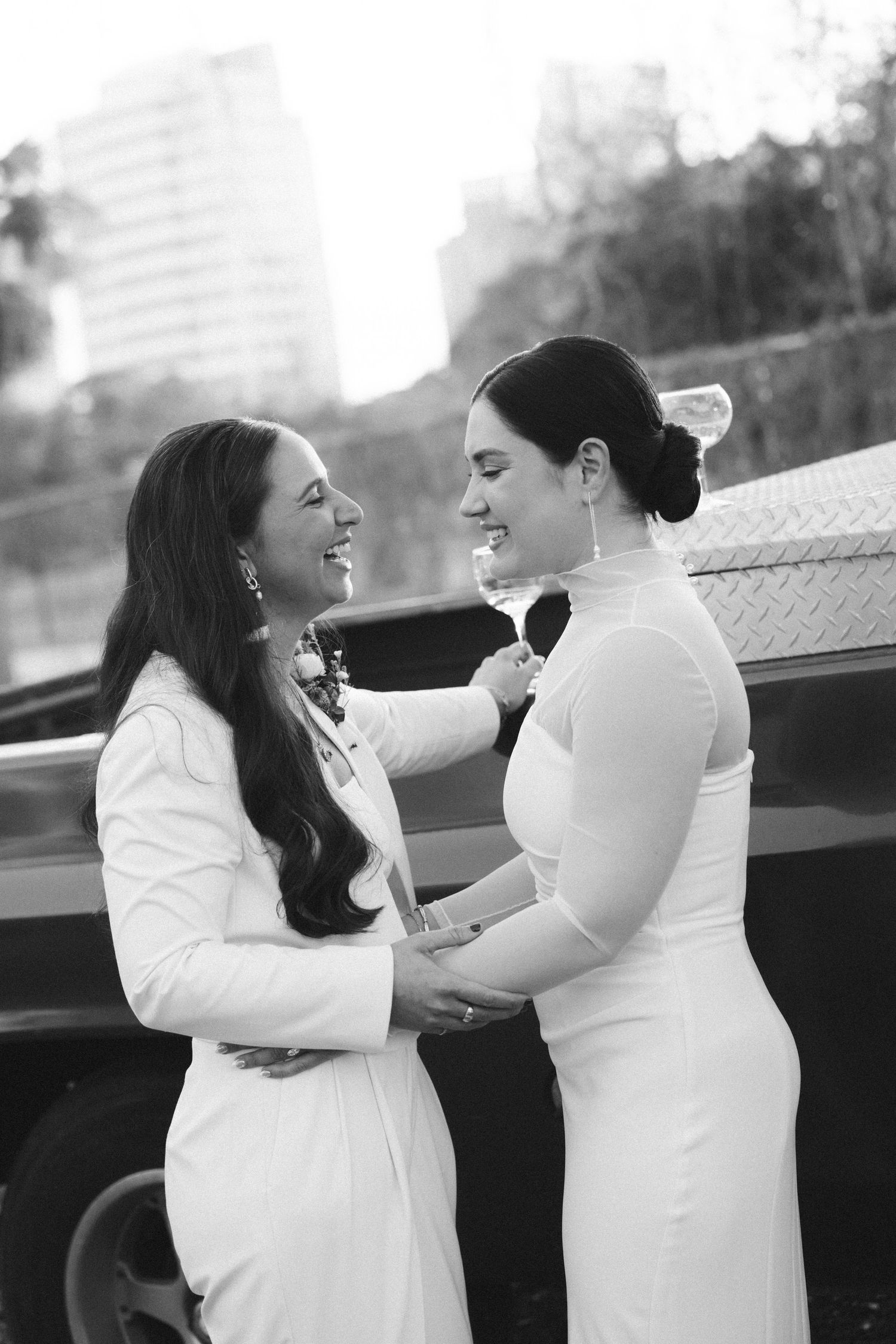 Two brides in white gowns share a joyful moment, smiling and holding hands in a black and white wedding photo.