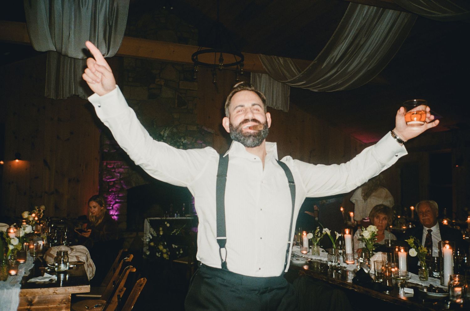 Bearded groom in suspenders celebrates with arms raised at candlelit wedding reception.